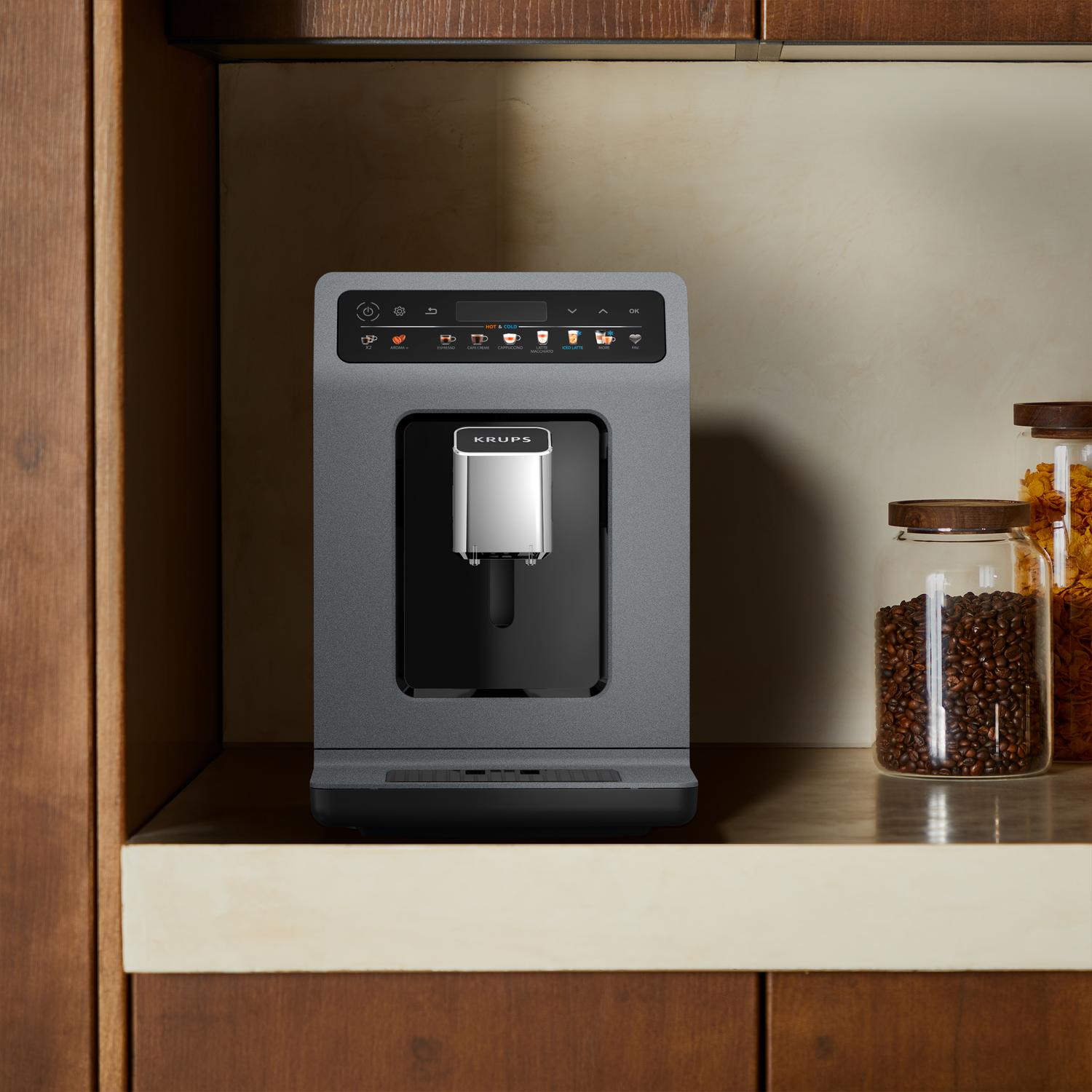 A grey Krups coffee machine sits on a shelf. Jars of coffee beans and grains are to the right. Wooden cabinetry surrounds it.