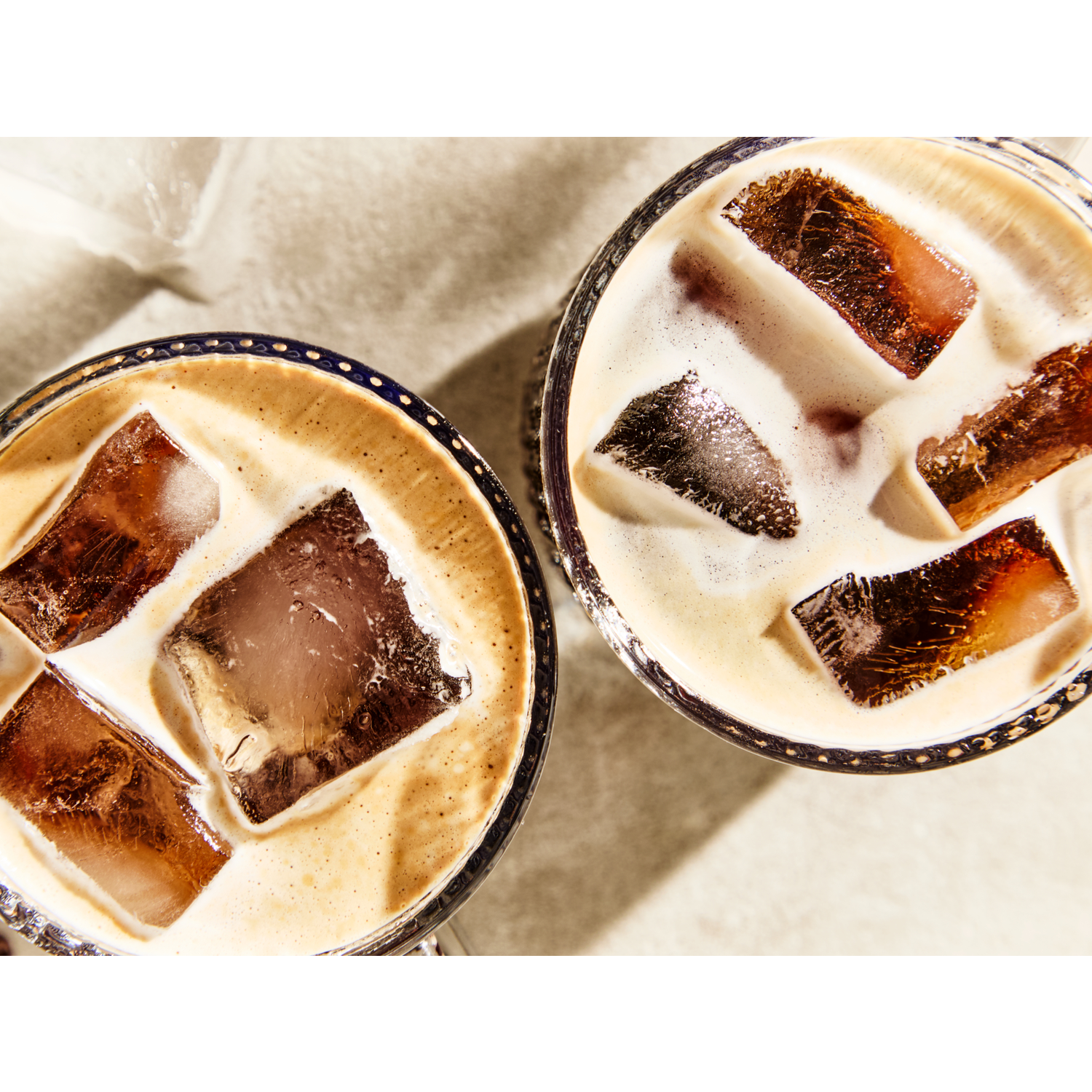 Two iced coffee drinks with large ice cubes. Coffee color is light brown with white foam. Drinks are in glass cups.