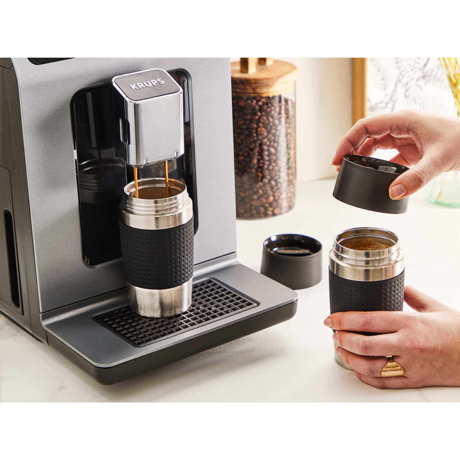 A coffee machine dispensing coffee into a travel mug. A hand is removing the lid of another mug.