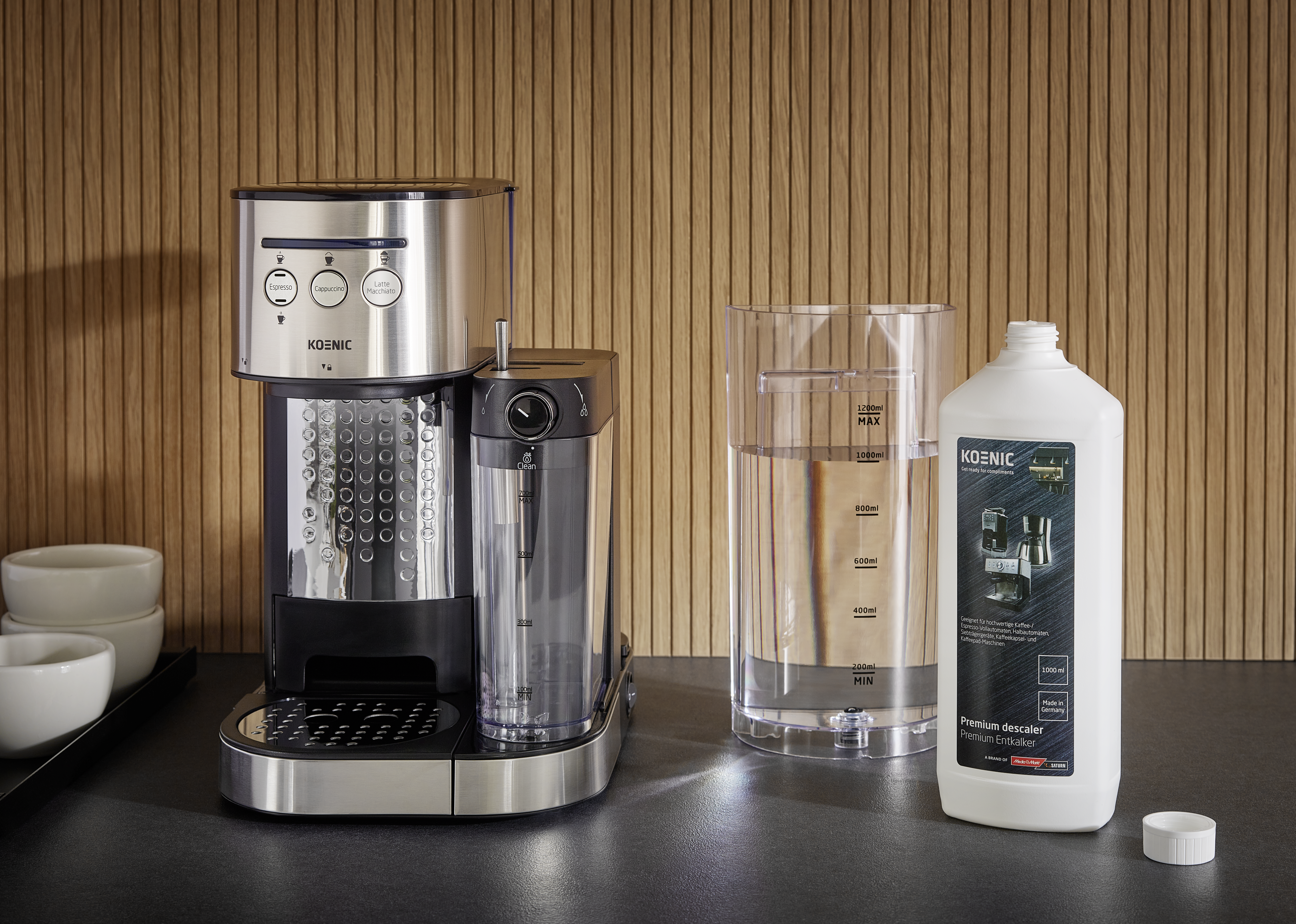 Kaffeemaschine, Wasserbehälter und Entkalkerflasche stehen auf dunkler Oberfläche. Hintergrund ist eine Holzwand.