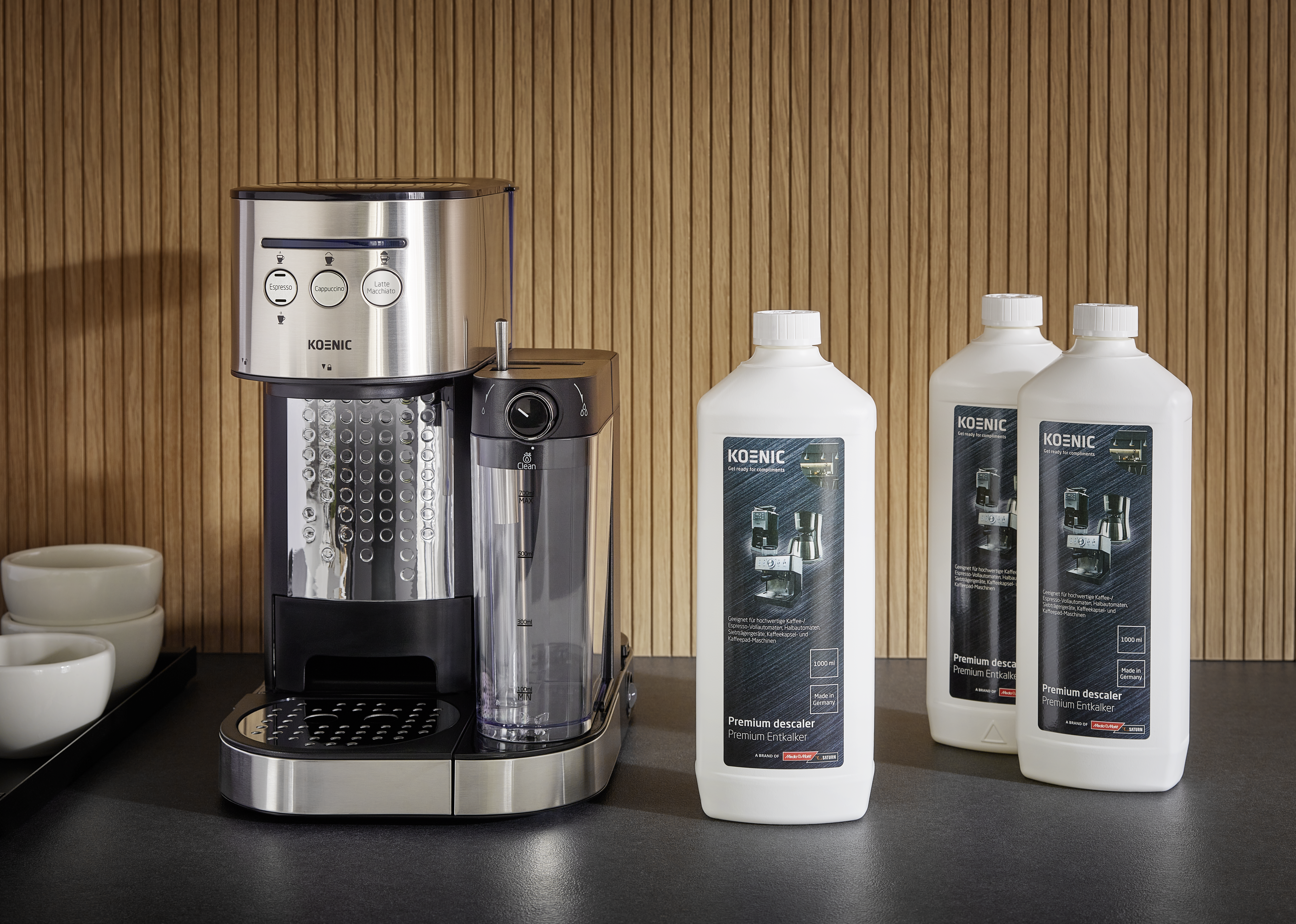 Eine Kaffeemaschine mit Entkalkerflaschen. Tassen befinden sich links. Holzpaneel-Hintergrund. Die Maschine hat drei Knöpfe.
