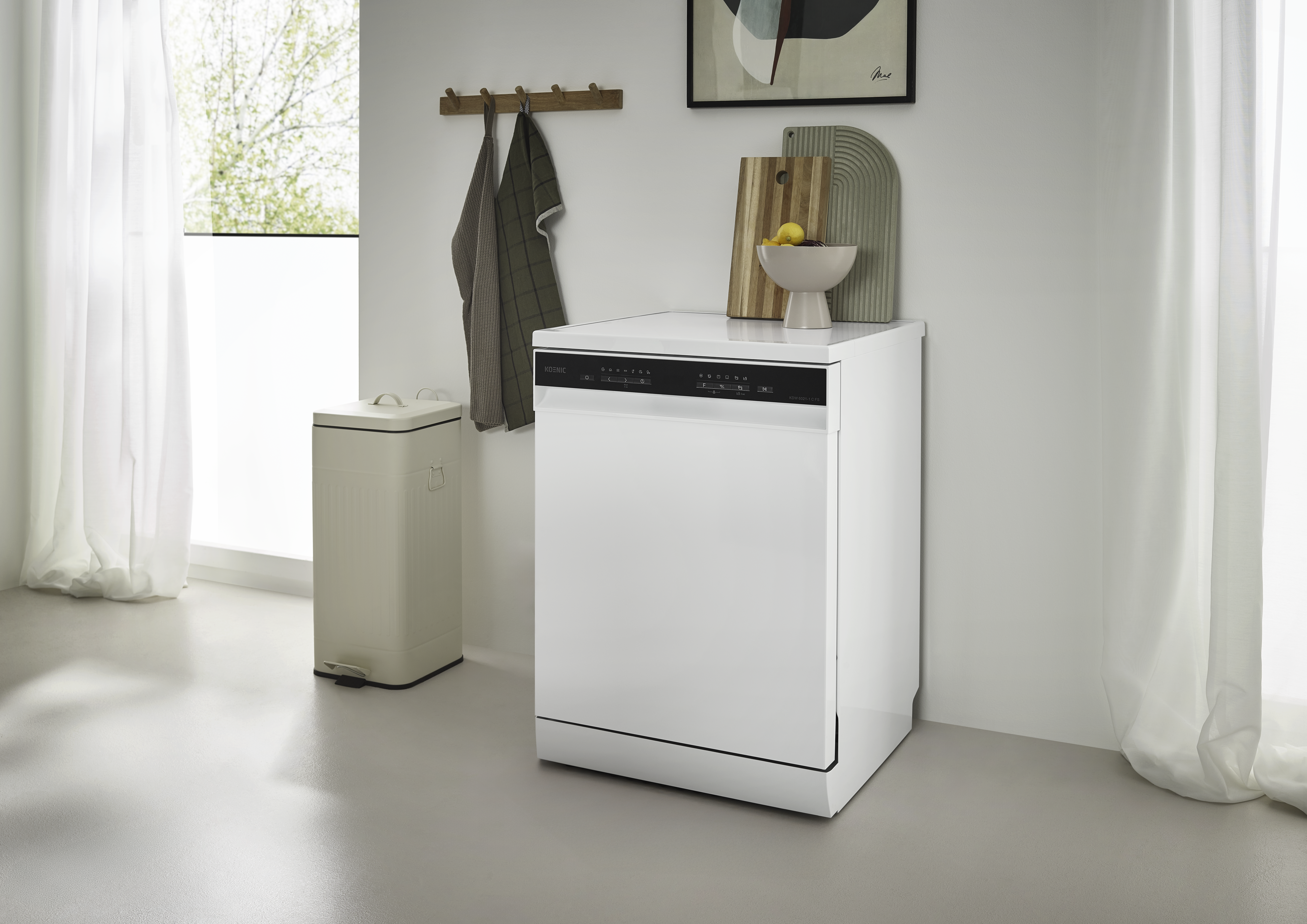 Ein weißer Geschirrspüler steht in der Nähe eines Fensters mit Vorhängen. Links steht ein beiger Mülleimer. Dekorative Gegenstände sind oben.