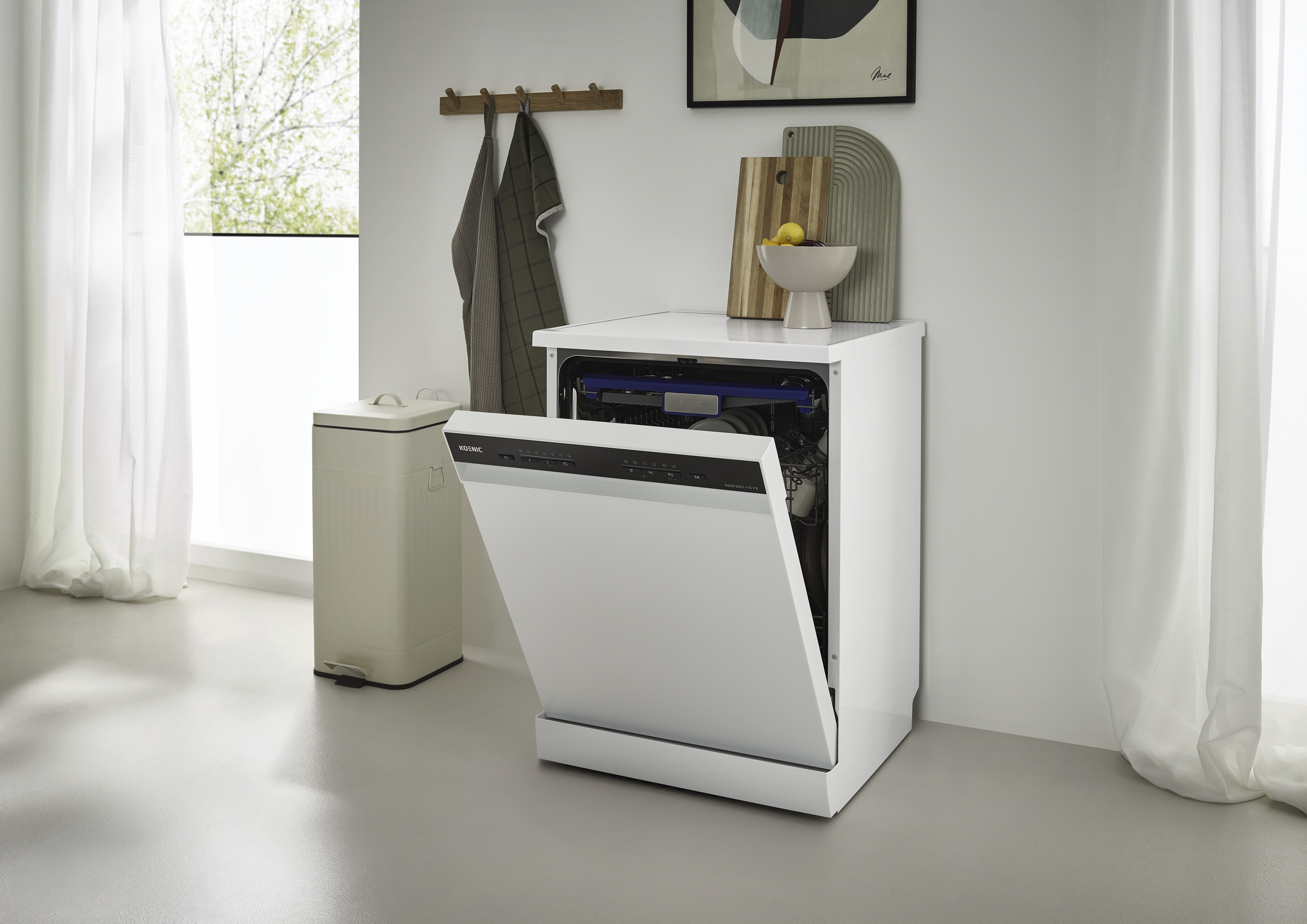 Eine weiße Spülmaschine mit offener Tür. Daneben ein Mülleimer und ein Fenster mit Vorhängen. Obenauf befinden sich Schneidebretter.