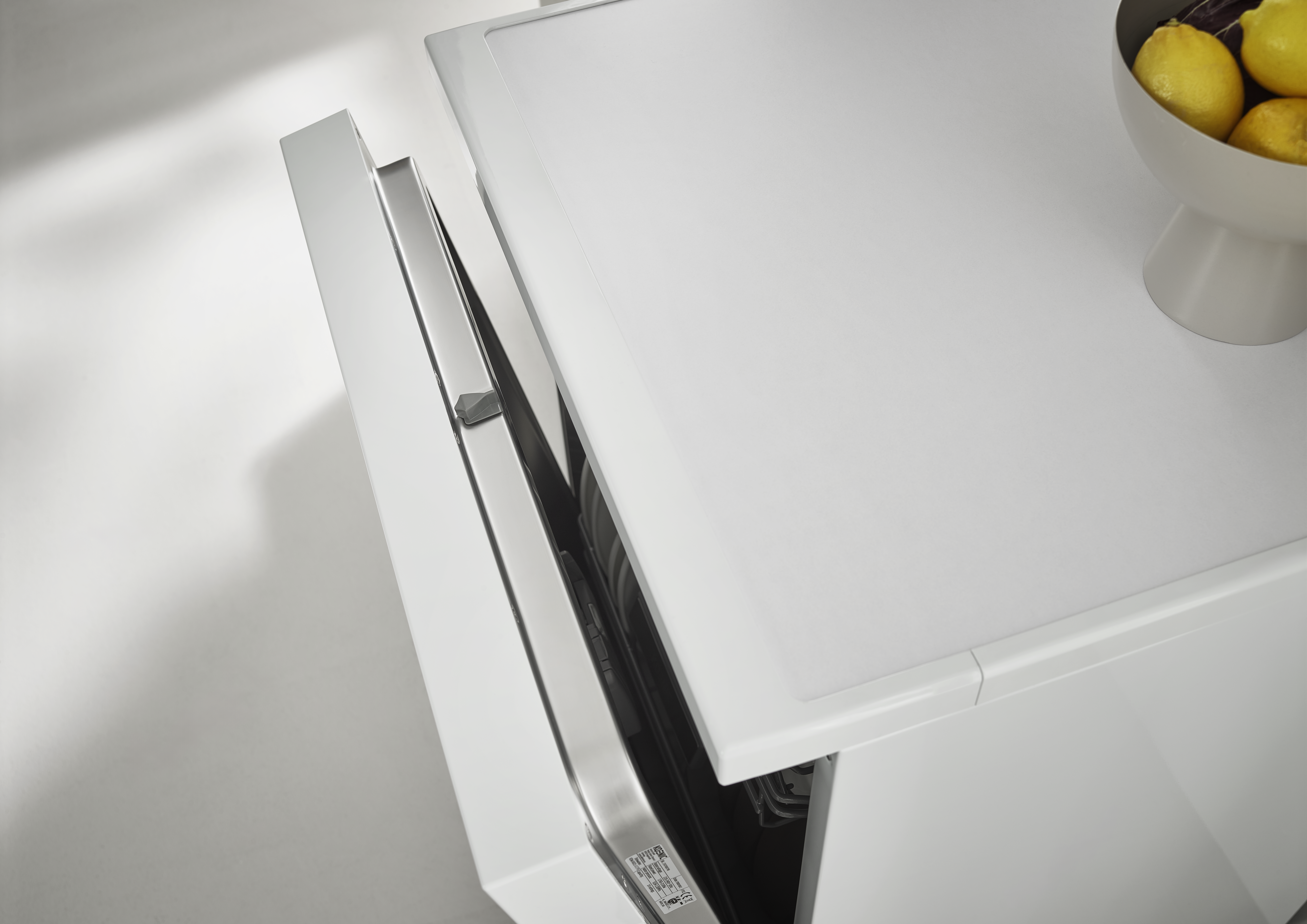 Eine weiße, teilweise geöffnete Spülmaschine mit Edelstahl-Innenraum. Eine Schale mit gelben Zitronen befindet sich rechts.