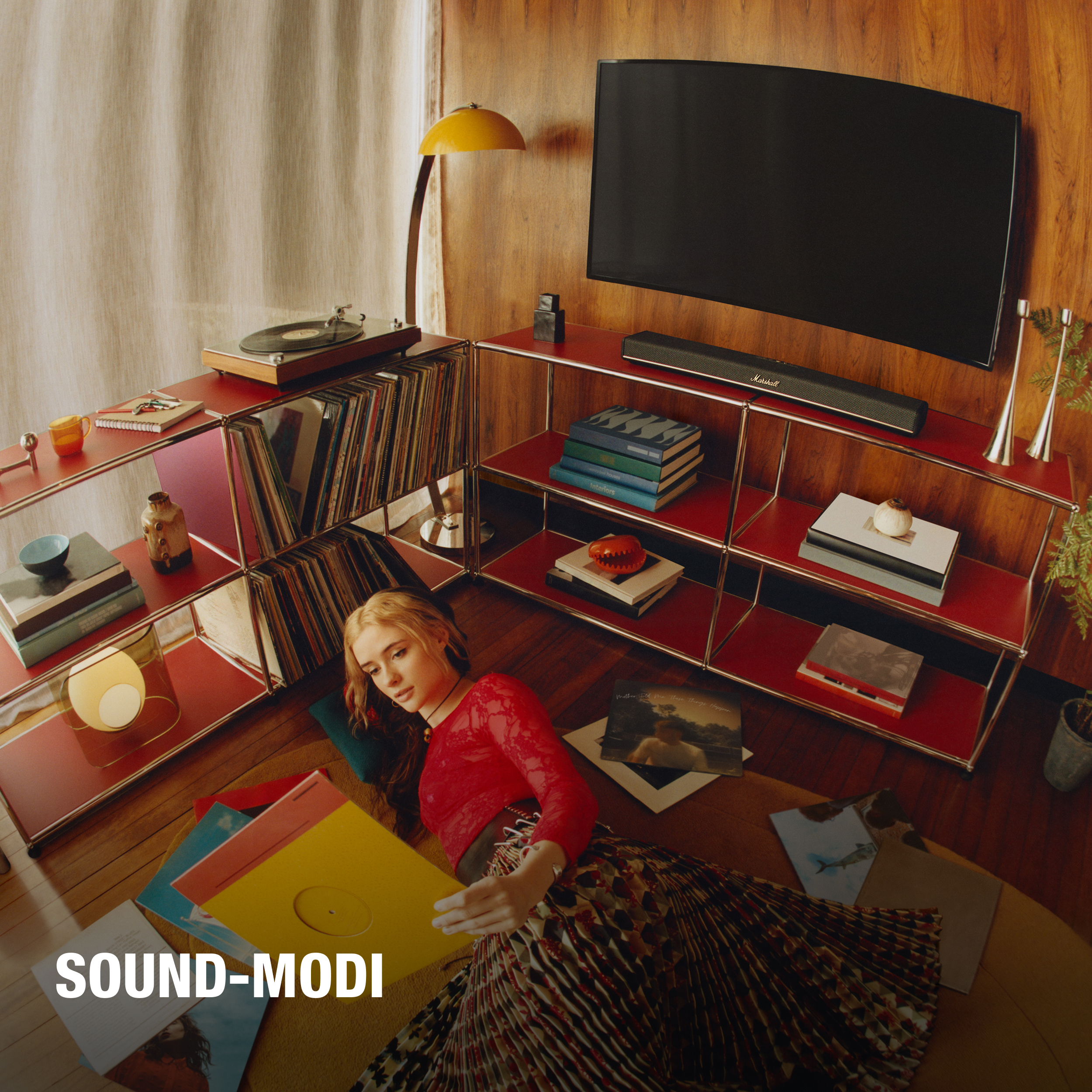 Eine Frau liegt mit Schallplatten auf dem Boden, rotes Oberteil. Regale mit Büchern, Plattenspieler und Lampe sichtbar.