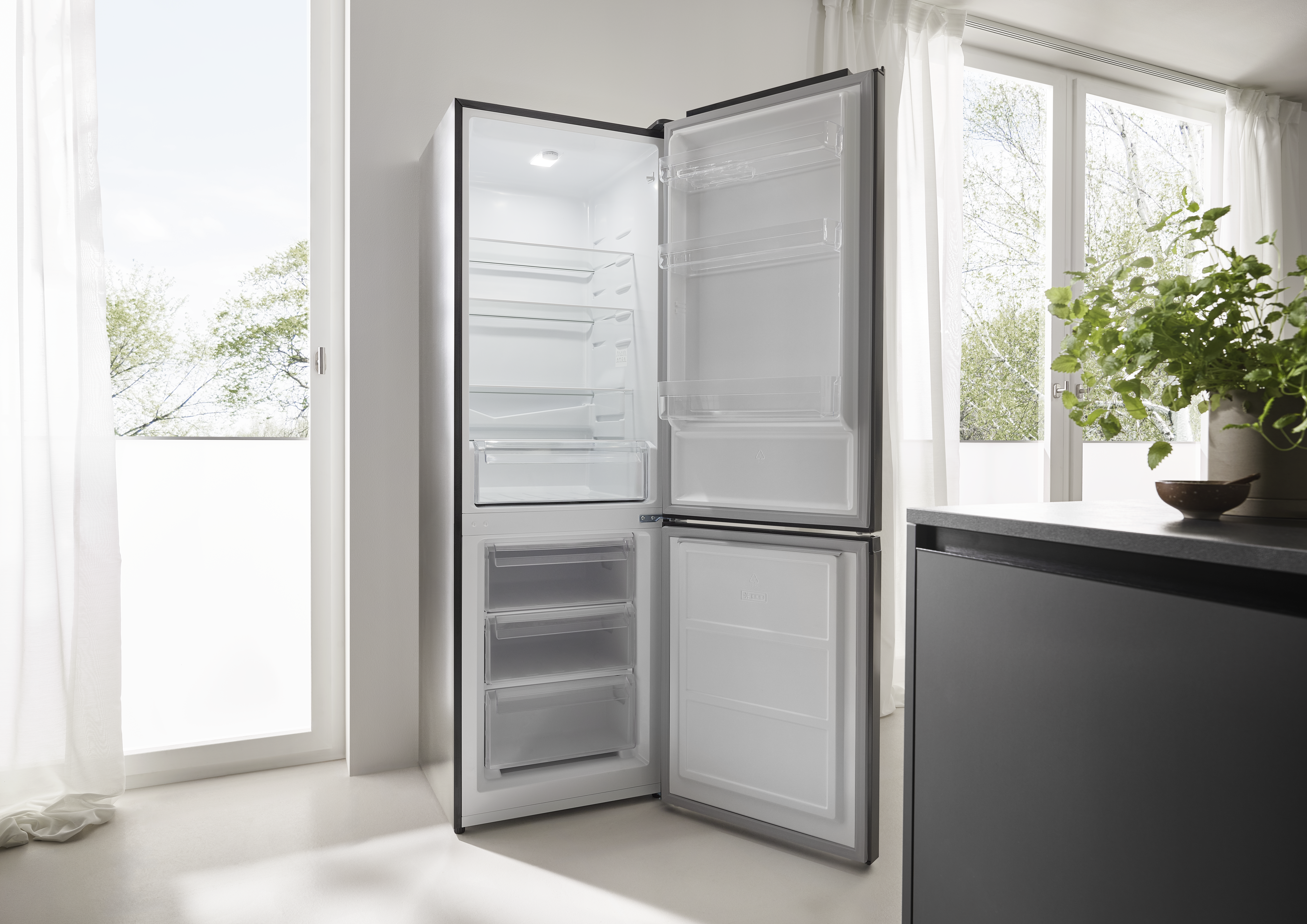 Ein offener Kühlschrank in einer hellen Küche. Die Kühlschranktüren sind geöffnet und zeigen Regale. Im Hintergrund sind ein Fenster und Pflanzen zu sehen.