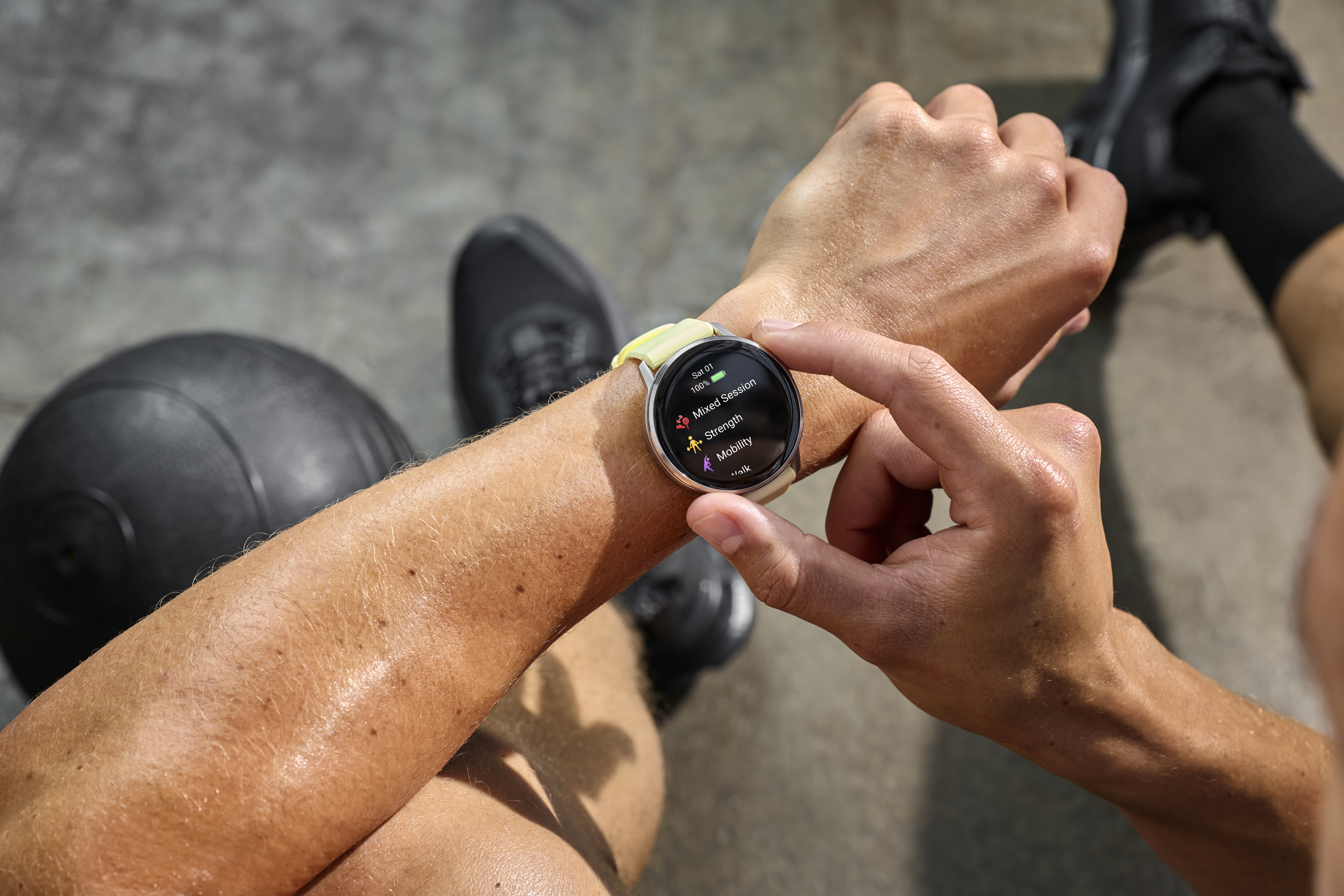 Eine Person überprüft eine Smartwatch mit Trainingsoptionen. Ein schwarzer Ball und Turnschuhe sind im Hintergrund, der Fokus liegt auf dem Handgelenk.