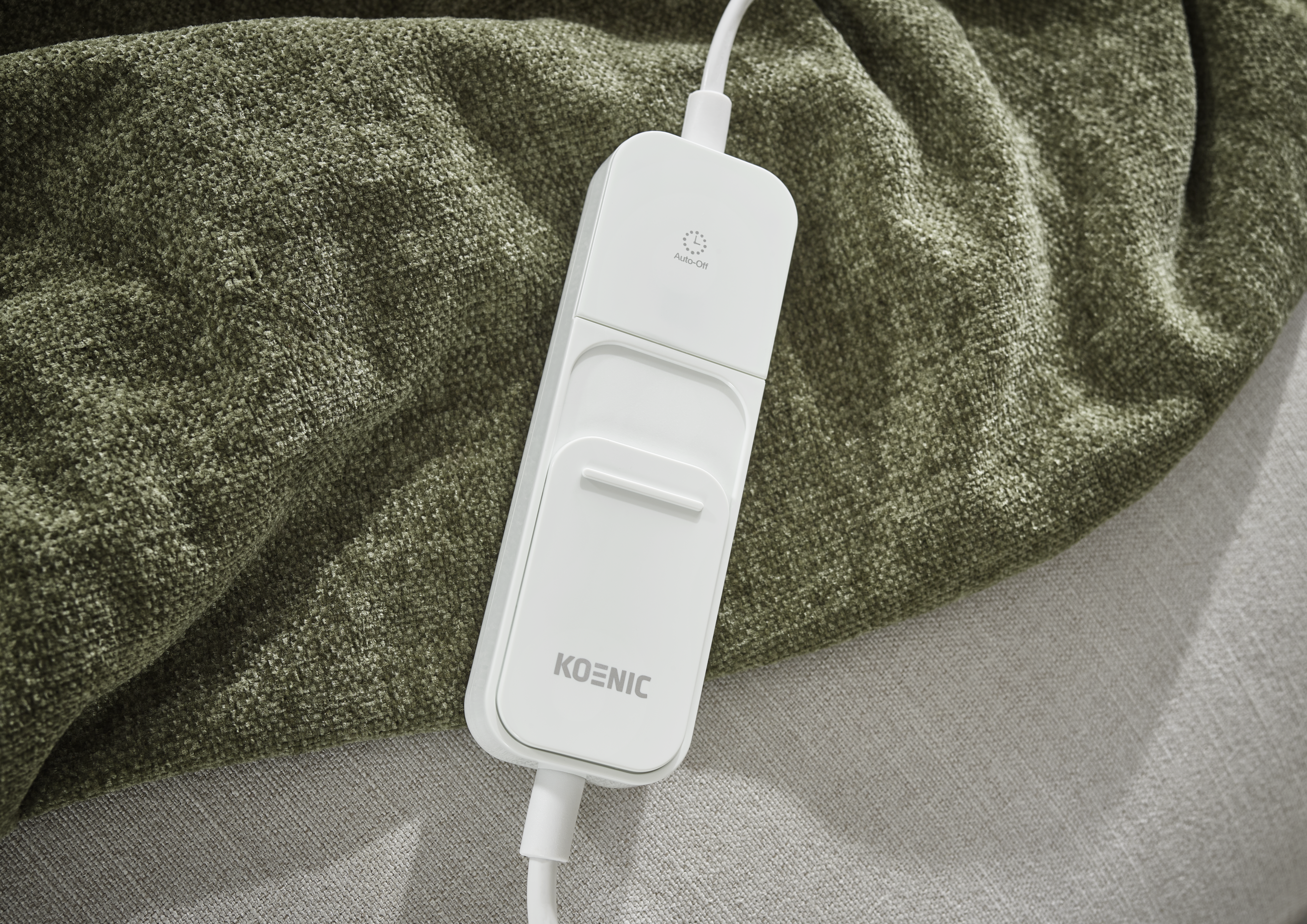 Eine weiße Koenic-Fernbedienung auf einer grün strukturierten Decke. Die Fernbedienung hat eine Auto-Off-Taste.