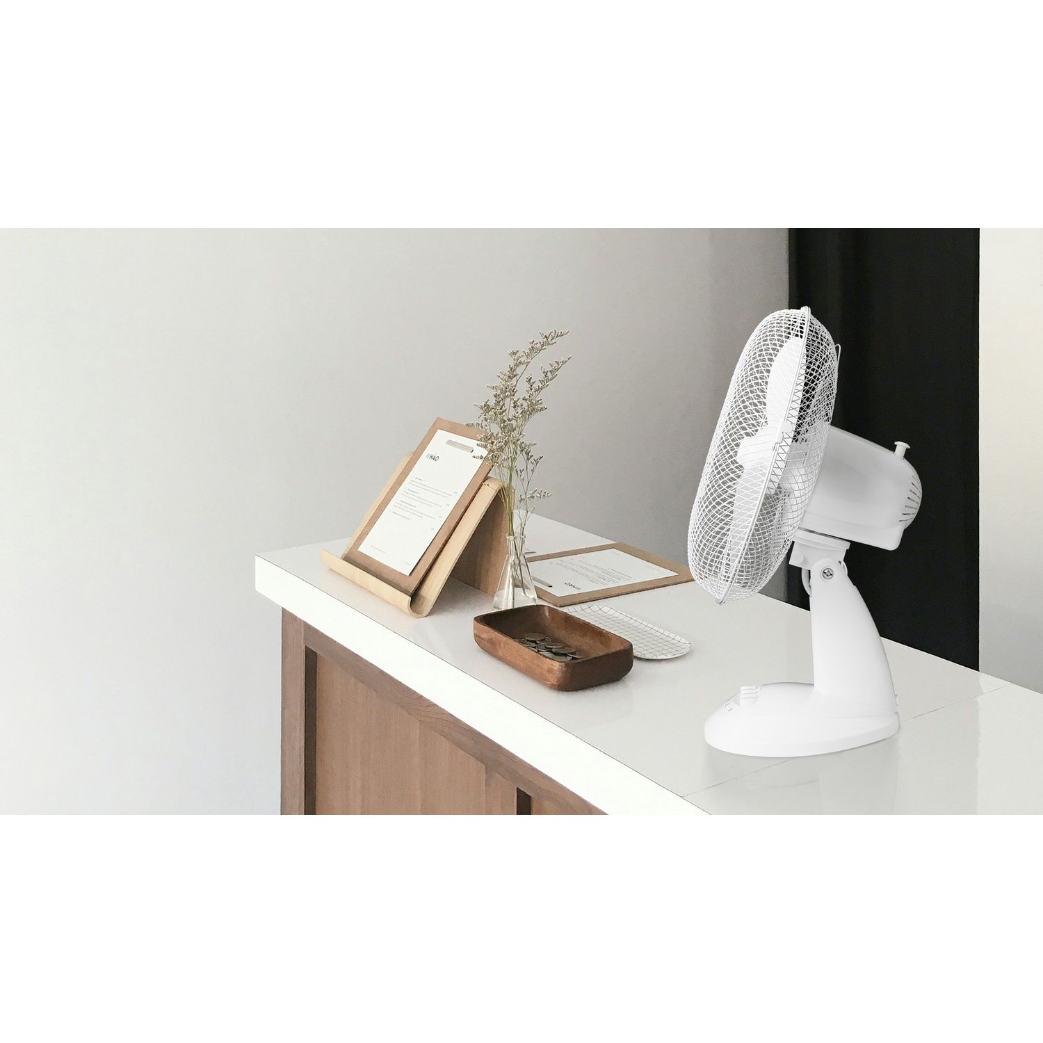 Un ventilatore da scrivania bianco, un vassoio di legno con monete e un menu incorniciato sono su un bancone bianco. Sono presenti anche fiori secchi e un menu.
