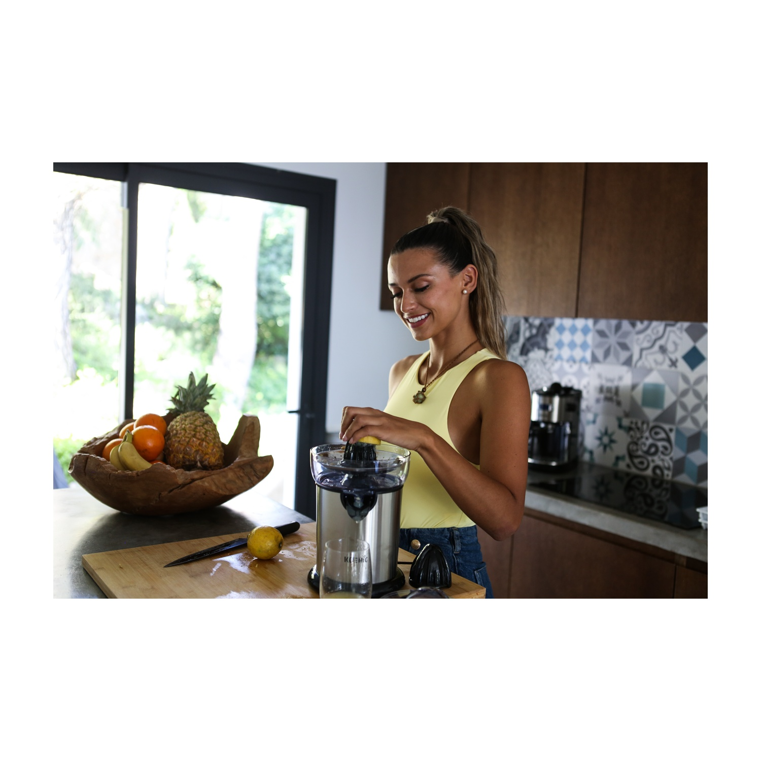 Una donna con un top giallo sorride mentre spremi un limone. Una ciotola di frutta è vicina. Cucina con spremiagrumi e finestra.