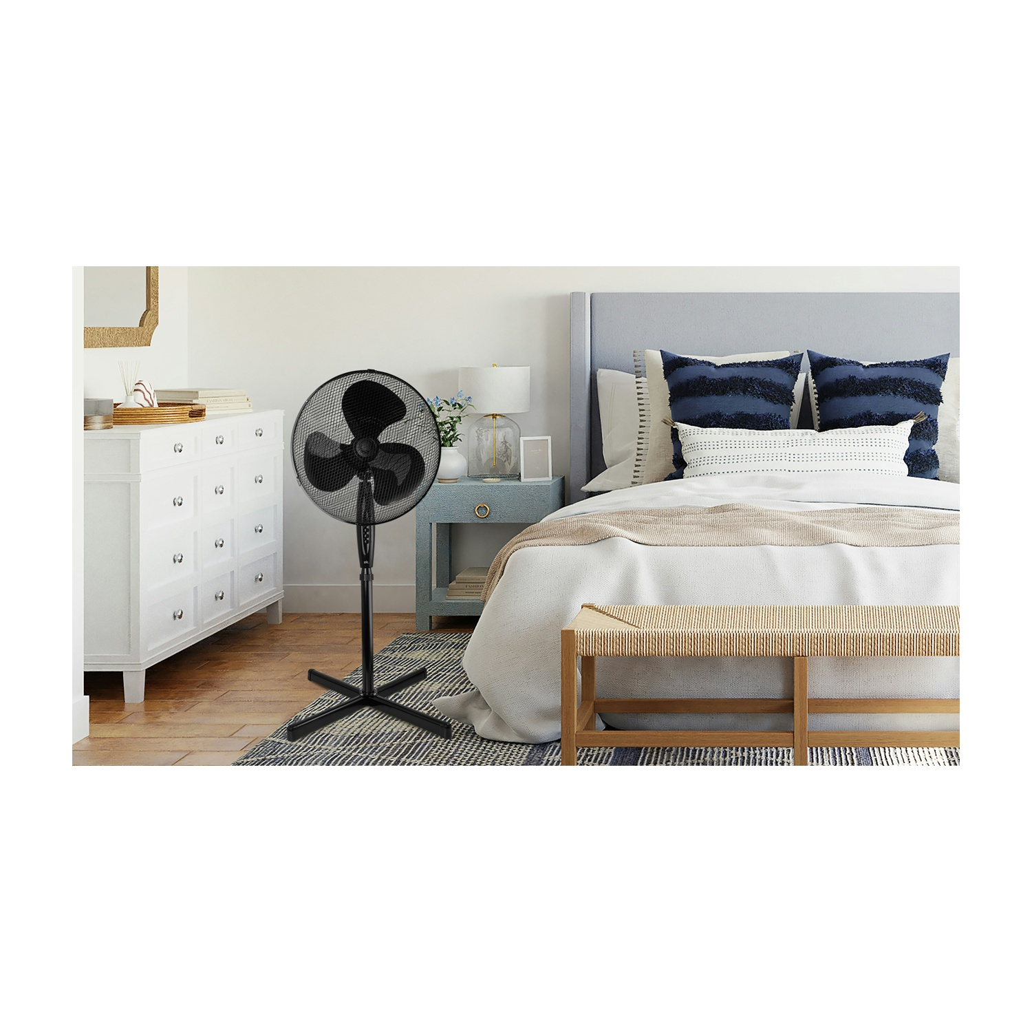 Un ventilatore nero in piedi in una camera da letto. Un letto con cuscini, una cassettiera bianca e una panca sono anche nella stanza.
