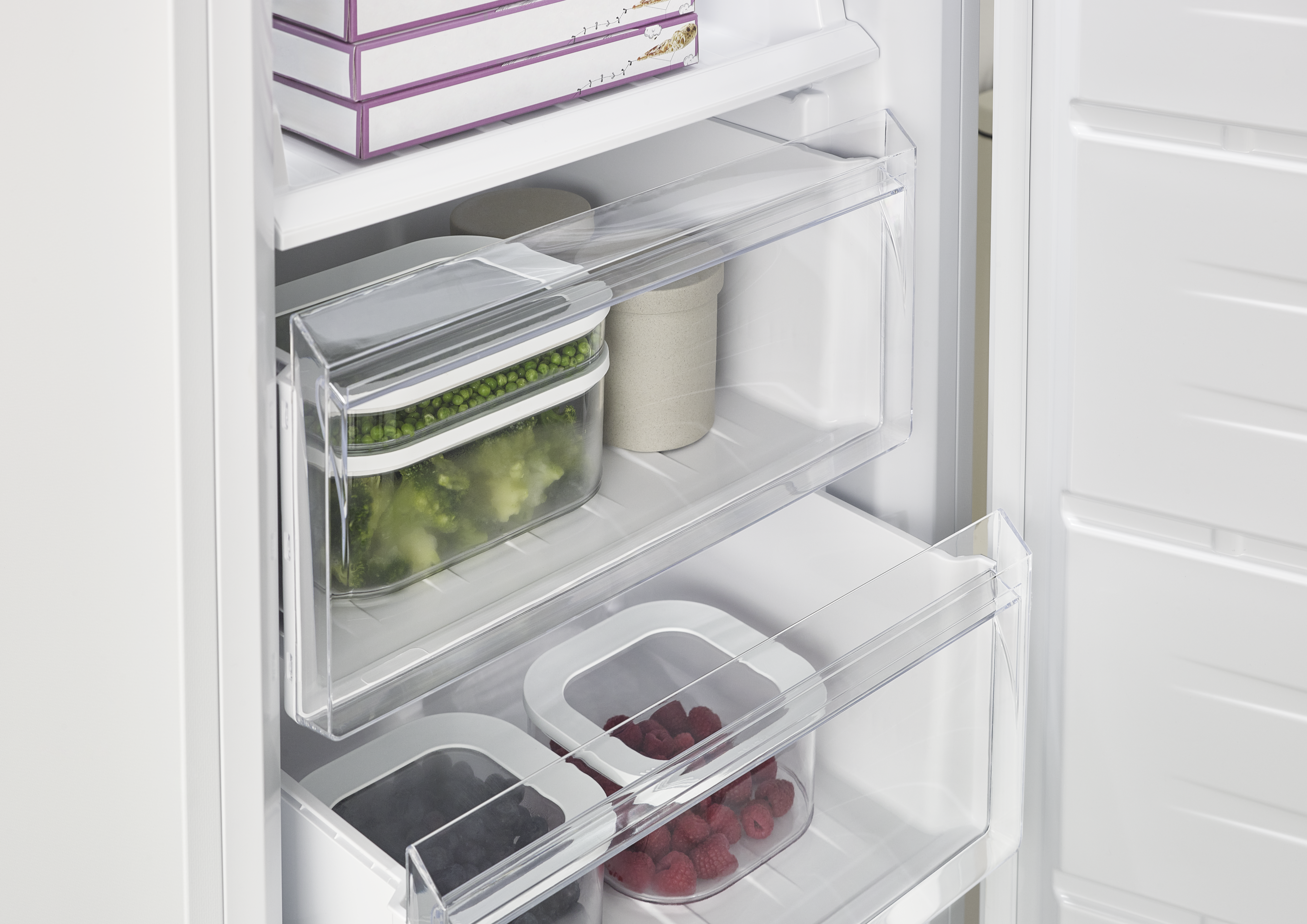 Offener Kühlschrank mit transparenten Schubladen. Erbsen, Himbeeren und Blaubeeren in Behältern. Weiße Regale und Türen. Einige Lebensmittelboxen.