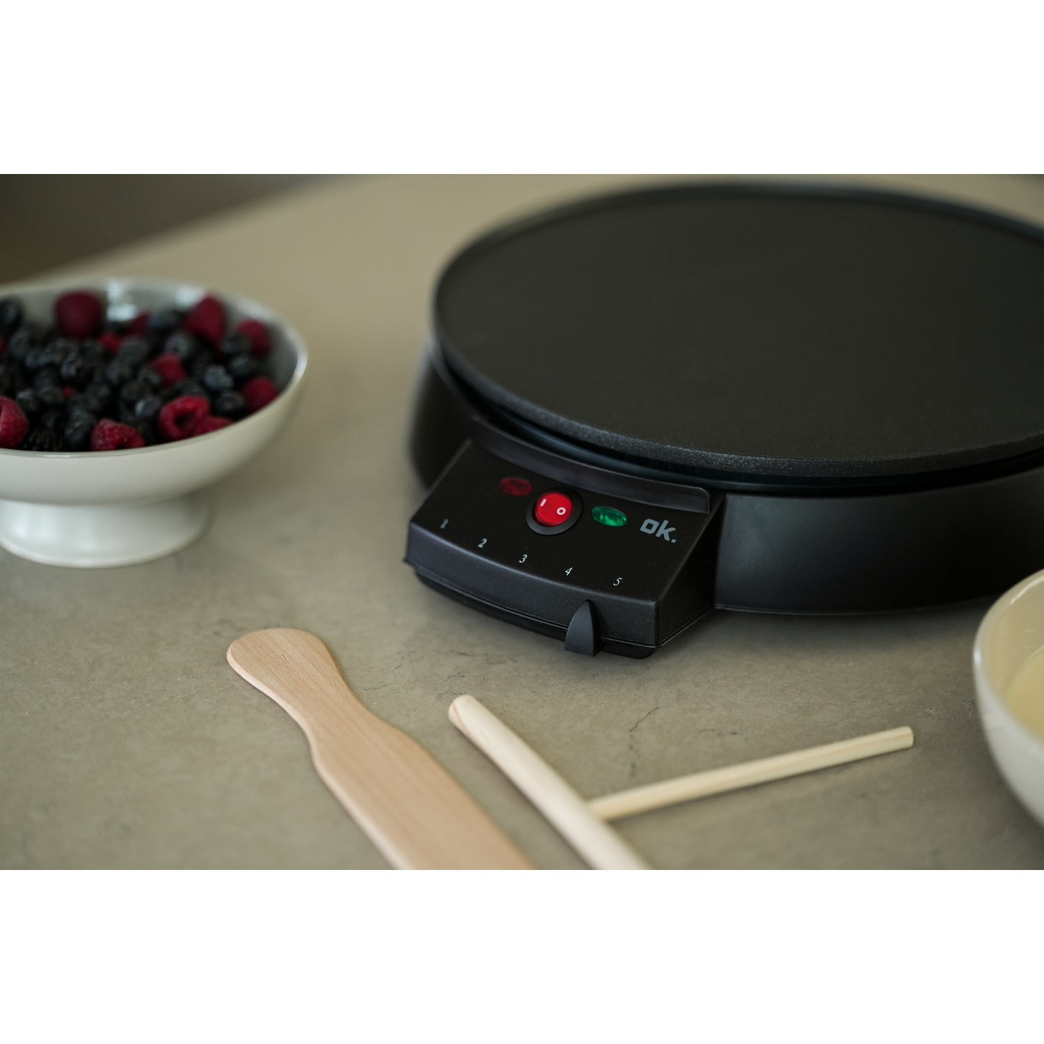 Una macchina per crêpes nera con pulsante di accensione rosso, una ciotola di bacche e utensili di legno su un piano grigio.
