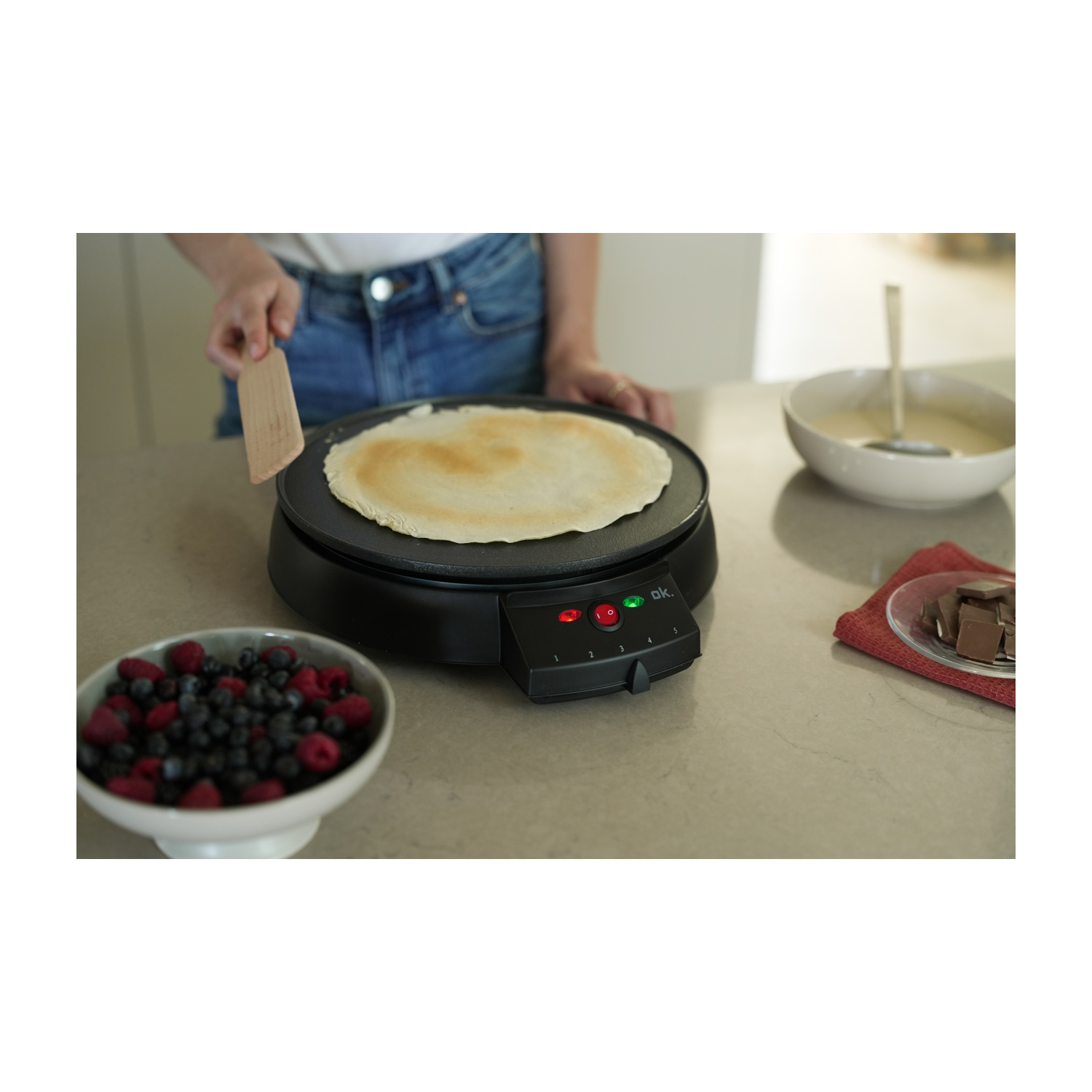Una persona prepara una crepe su una piastra nera. Ci sono bacche, pastella e cioccolato nelle vicinanze. Viene utilizzata una spatola di legno.
