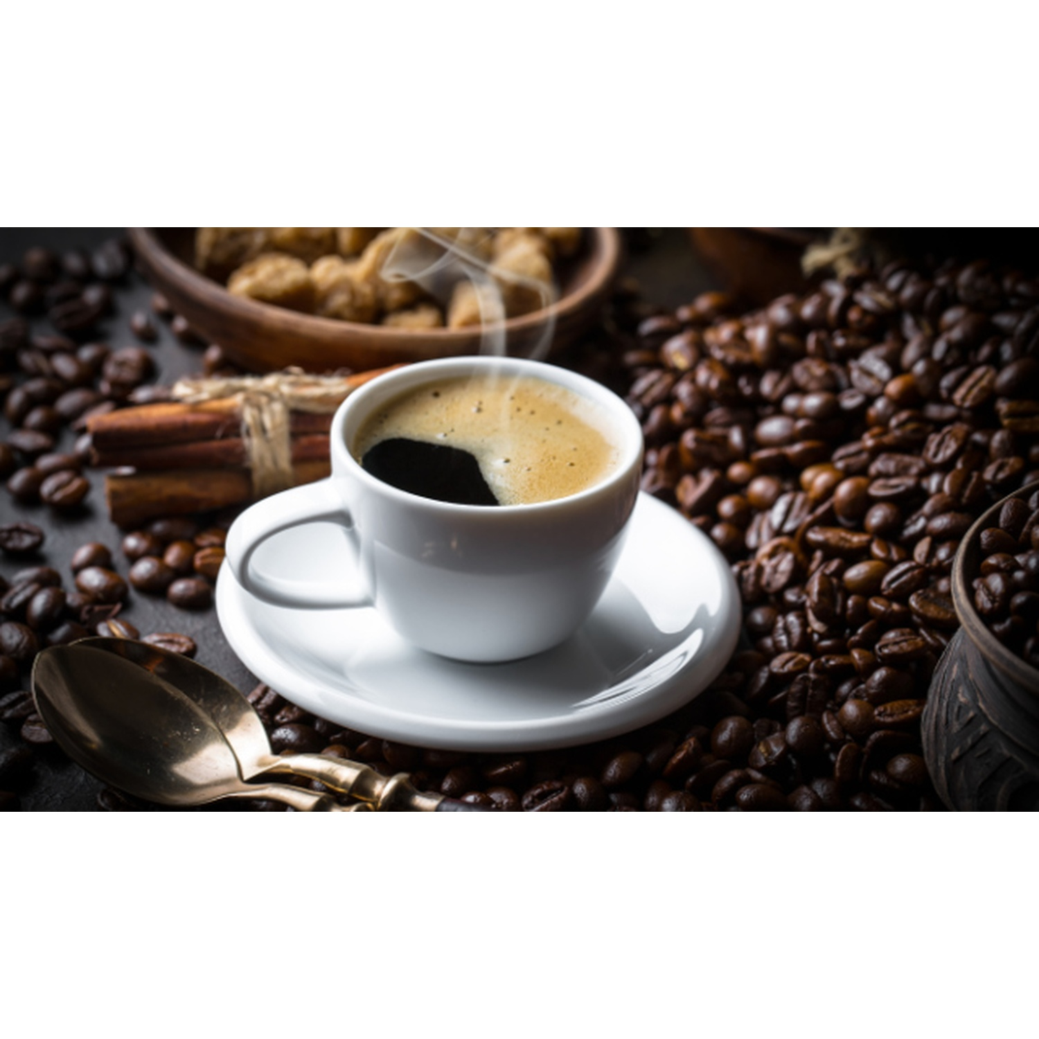 Una tazza bianca di caffè su un piattino, circondata da chicchi di caffè, bastoncini di cannella e zollette di zucchero in una ciotola di legno.