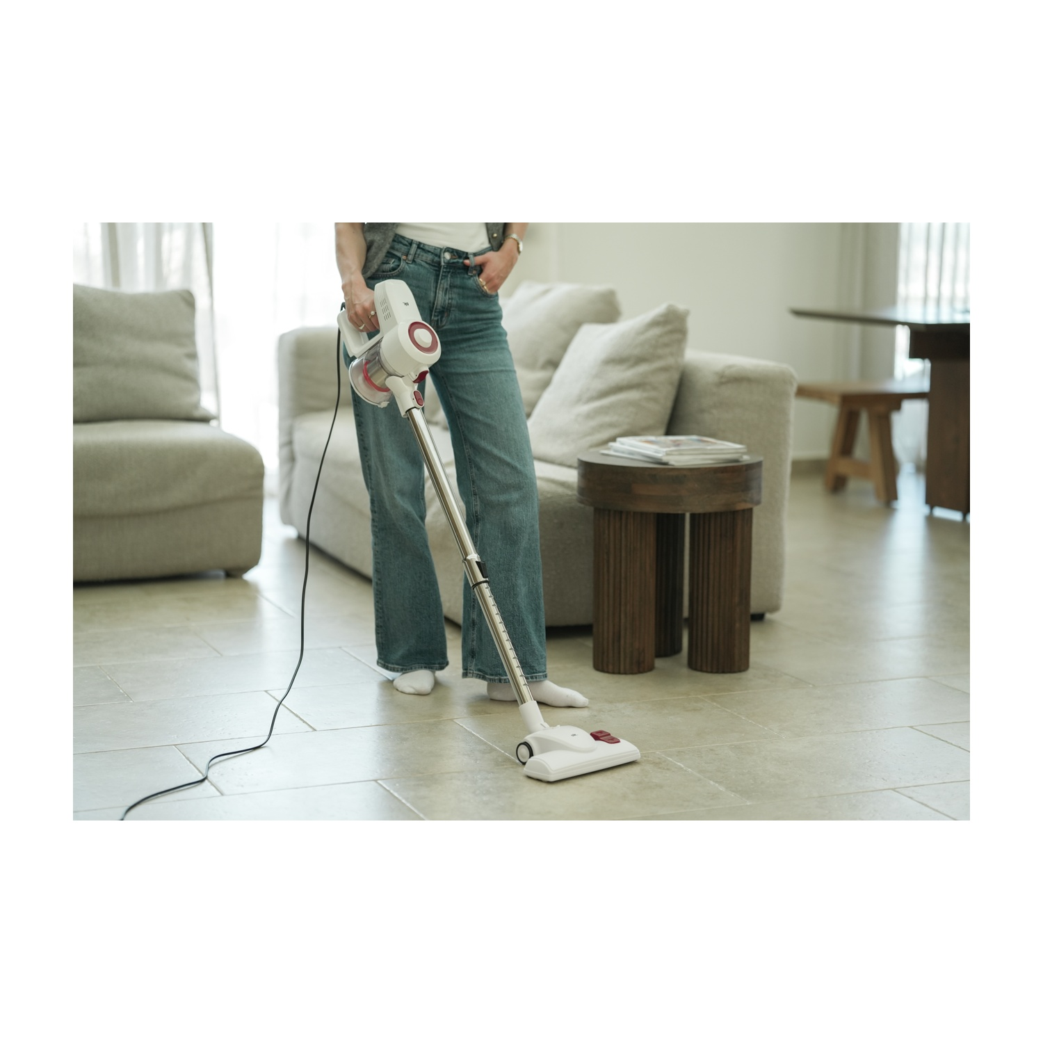 Una donna in jeans passa l'aspirapolvere su un pavimento piastrellato beige con un aspirapolvere bianco e rosso. Sullo sfondo un divano e un tavolo di legno.