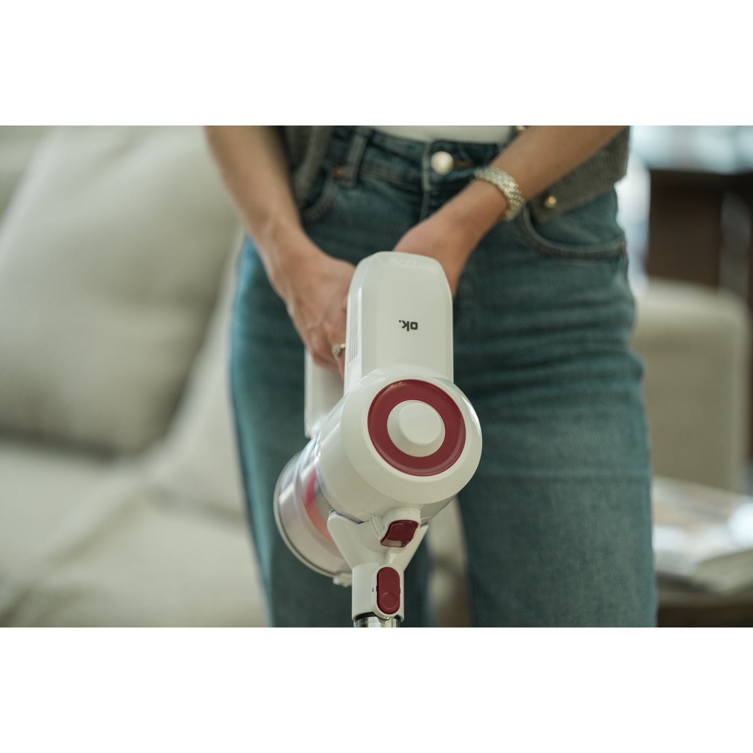 Una persona tiene in mano un aspirapolvere senza fili bianco e rosso. Indossa jeans e una giacca grigia. Un divano è sullo sfondo.