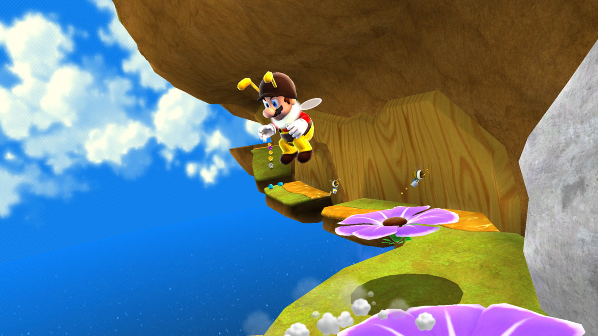 Mario in costume da ape salta sulle piattaforme. Un grande fiore in primo piano. Lo sfondo mostra cielo blu con nuvole.