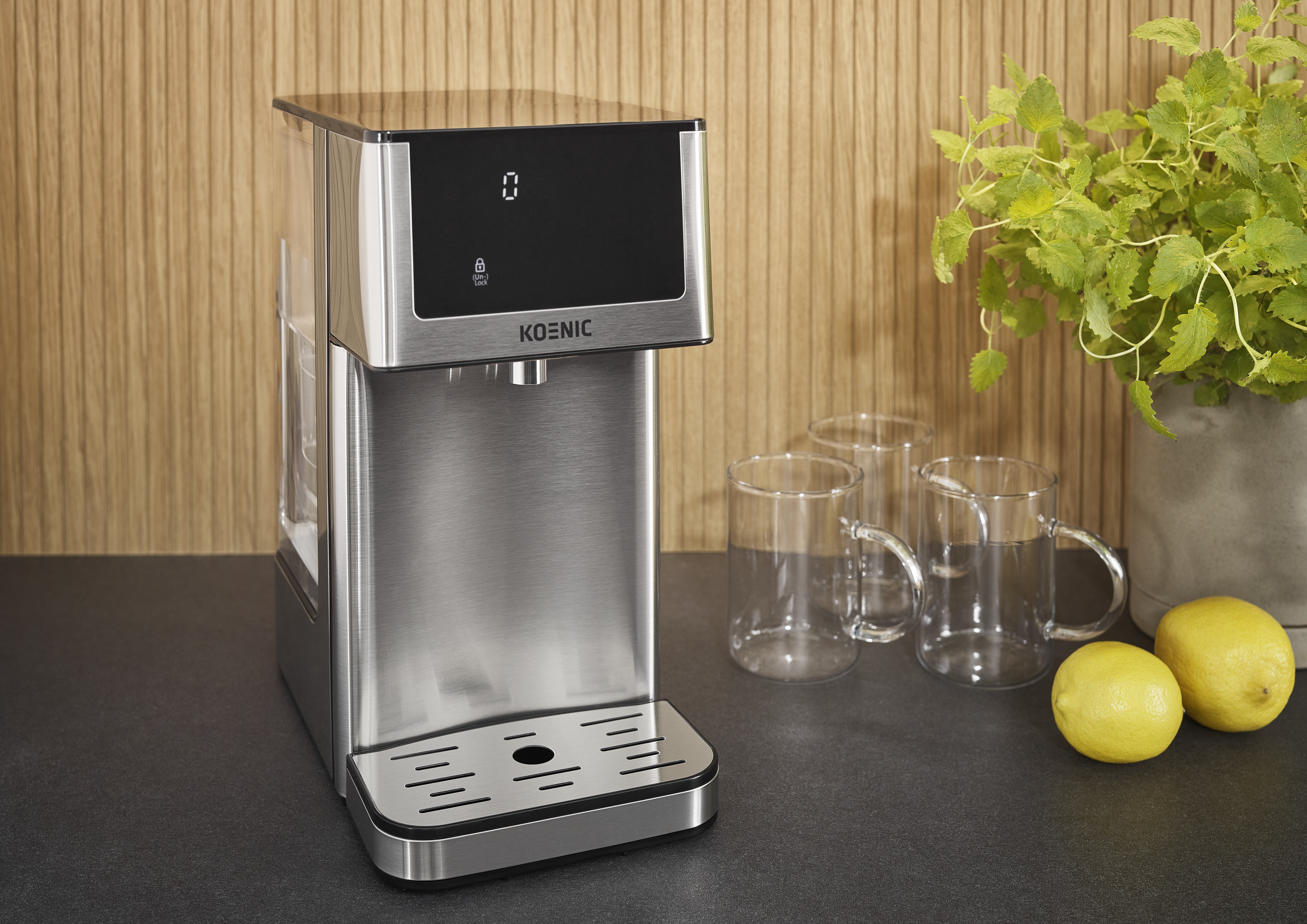Ein Edelstahl-Wasserspender mit drei Glasbechern, zwei Zitronen und einer Pflanze. Der Hintergrund ist eine Holzverkleidung.