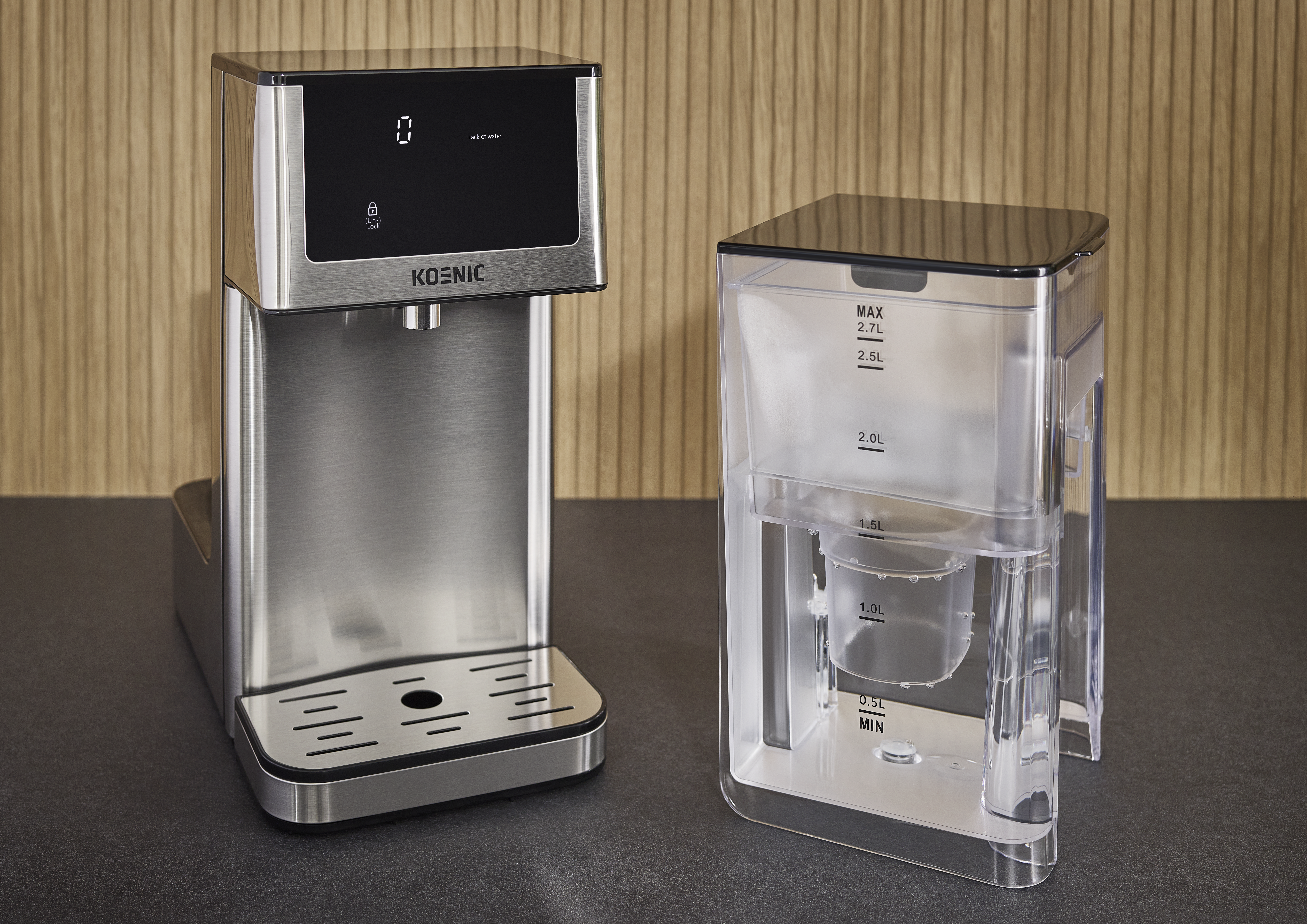 Ein silberner Wasserspender mit schwarzem Display und ein transparenter Wassertank daneben auf dunkler Oberfläche.