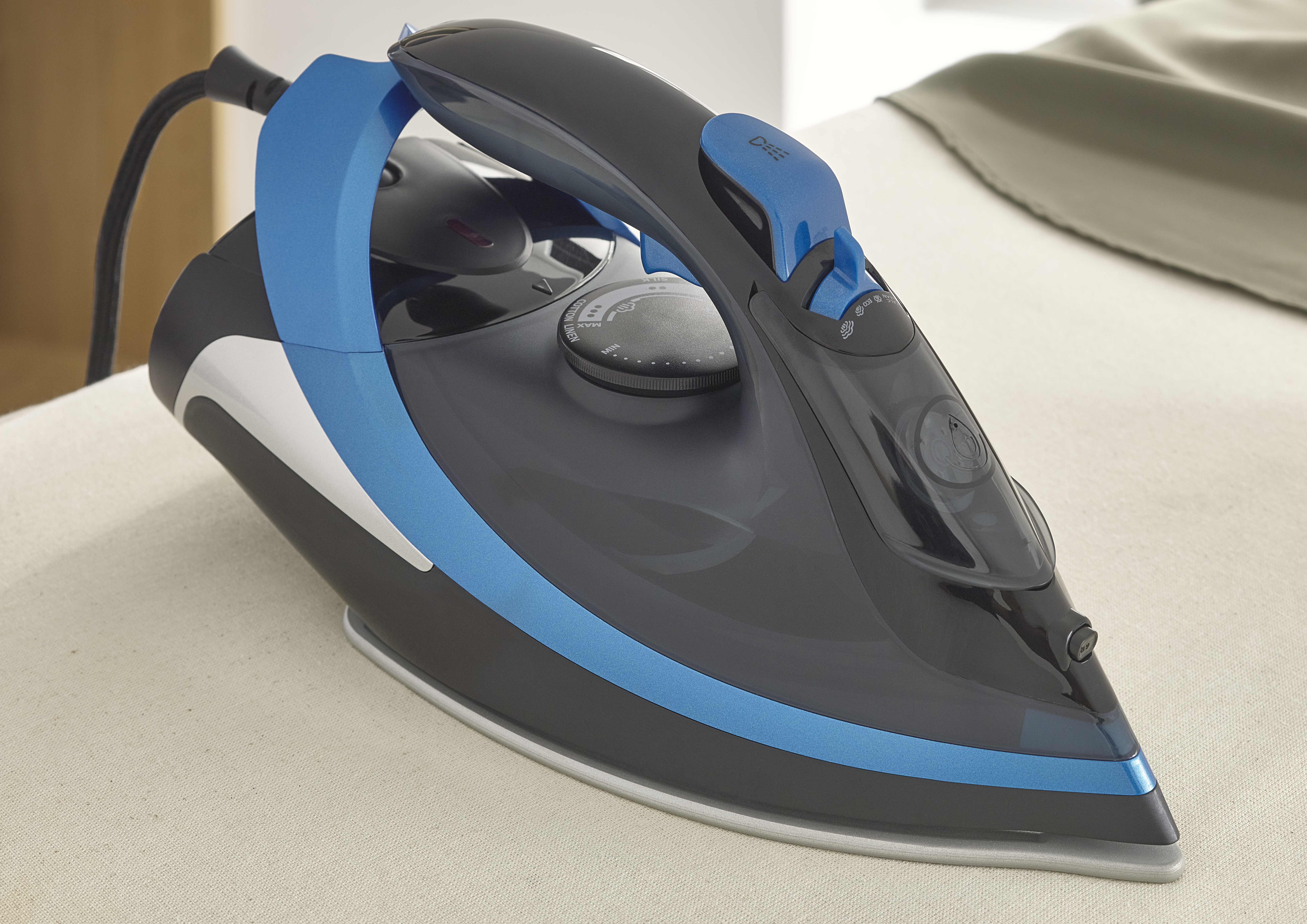 Ein schwarz-blaues Bügeleisen auf einer beigefarbenen Oberfläche. Im Hintergrund befindet sich ein grüner Stoff. Das Bügeleisen hat ein schwarzes Kabel.