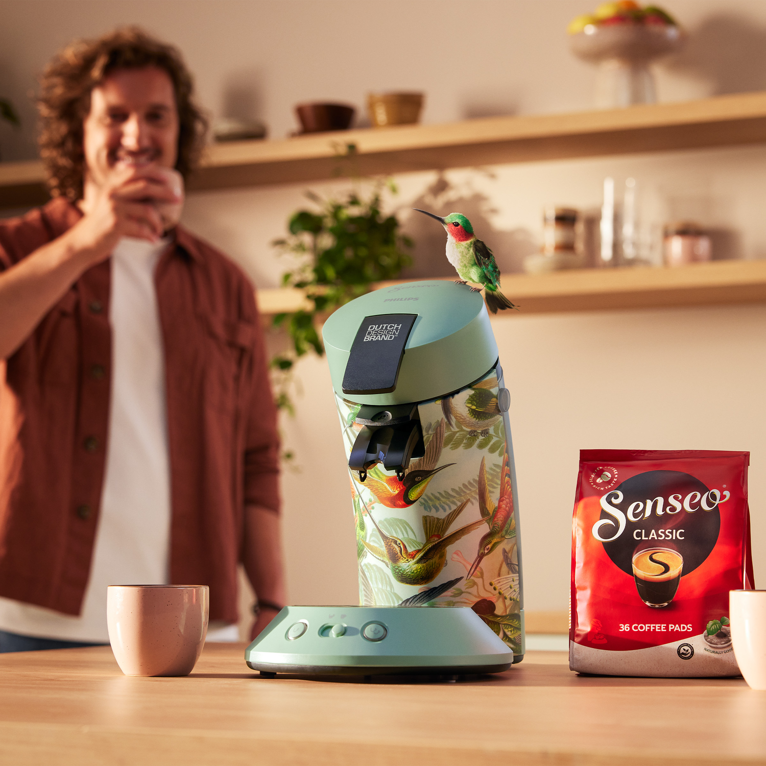 A man is drinking coffee, a coffee machine with hummingbird design, a bag of coffee pads, and a hummingbird.