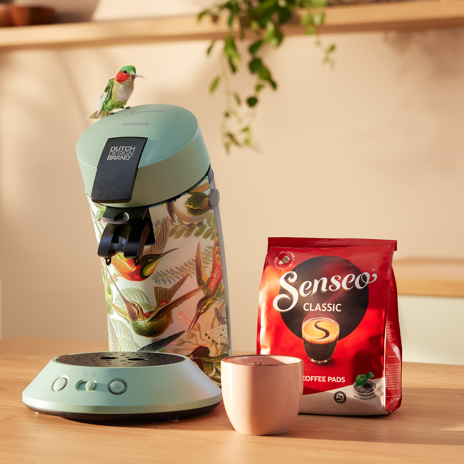 A Senseo coffee machine with a hummingbird perched on it. A bag of Senseo coffee pads and a small cup sit nearby on a wooden table.