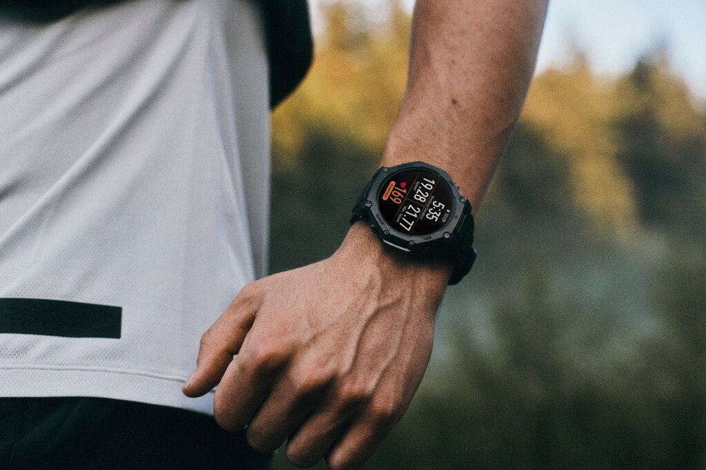 Brazo de una persona con un reloj inteligente negro. El reloj muestra la hora y la frecuencia cardíaca. El fondo son árboles borrosos.