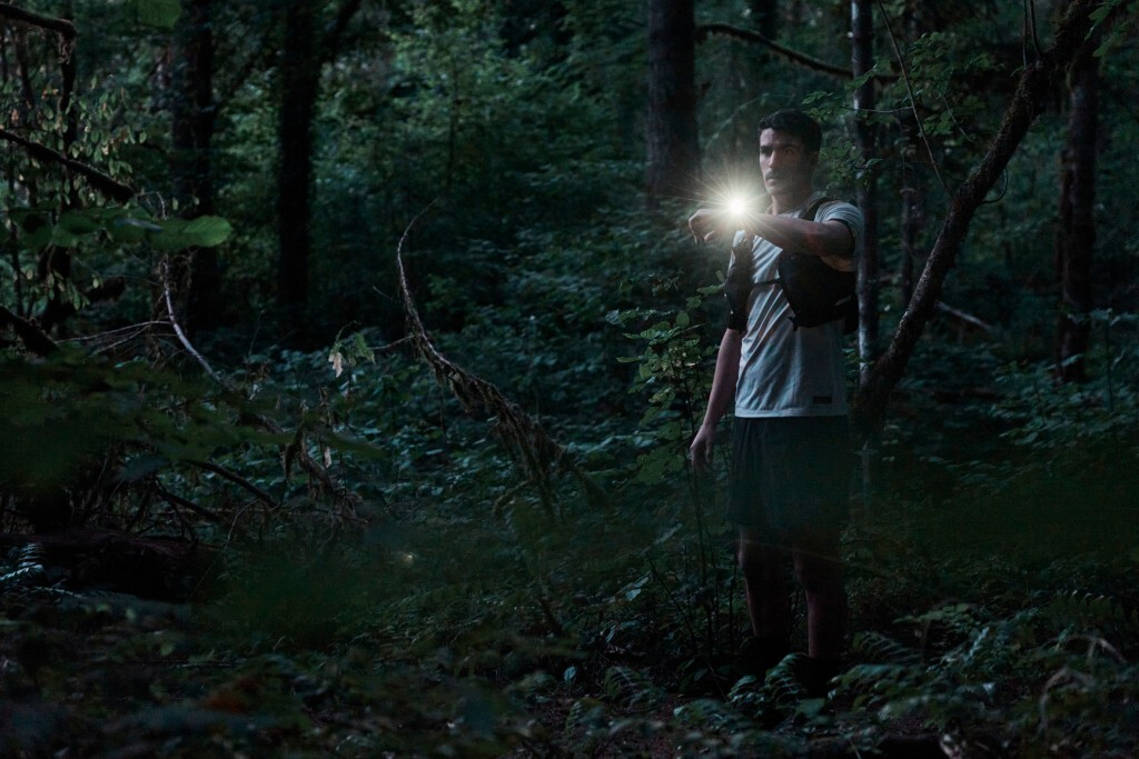 Un hombre en un bosque sostiene una luz brillante. Lleva camisa y pantalón corto. El follaje verde oscuro lo rodea.