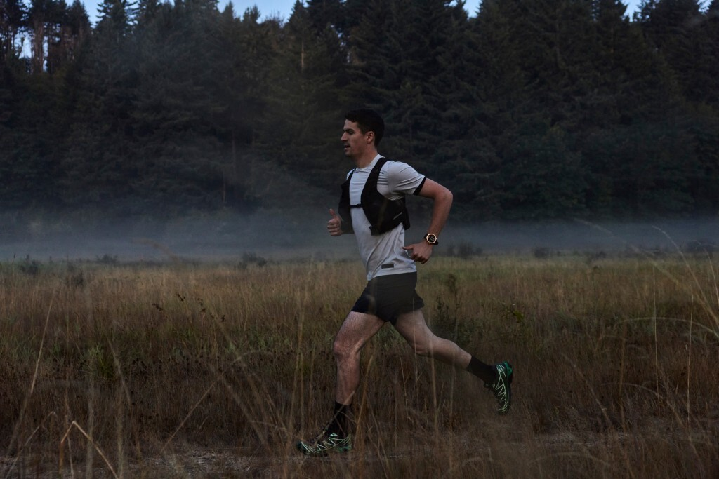 Un hombre corre por un campo con hierba alta. Lleva una camisa blanca, pantalones cortos negros y un chaleco. Hay árboles al fondo.