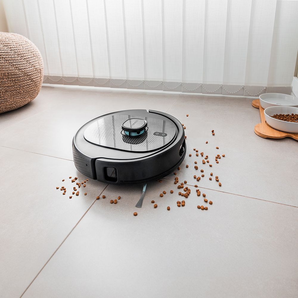 Una aspiradora robot negra limpia un suelo embaldosado con comida para mascotas esparcida. Un puf tejido y persianas venecianas en el fondo.