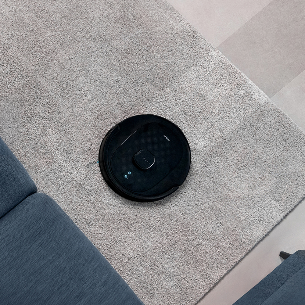Una aspiradora robótica negra sobre una alfombra gris. Un sofá azul es visible a la izquierda. El fondo muestra un suelo de baldosas.
