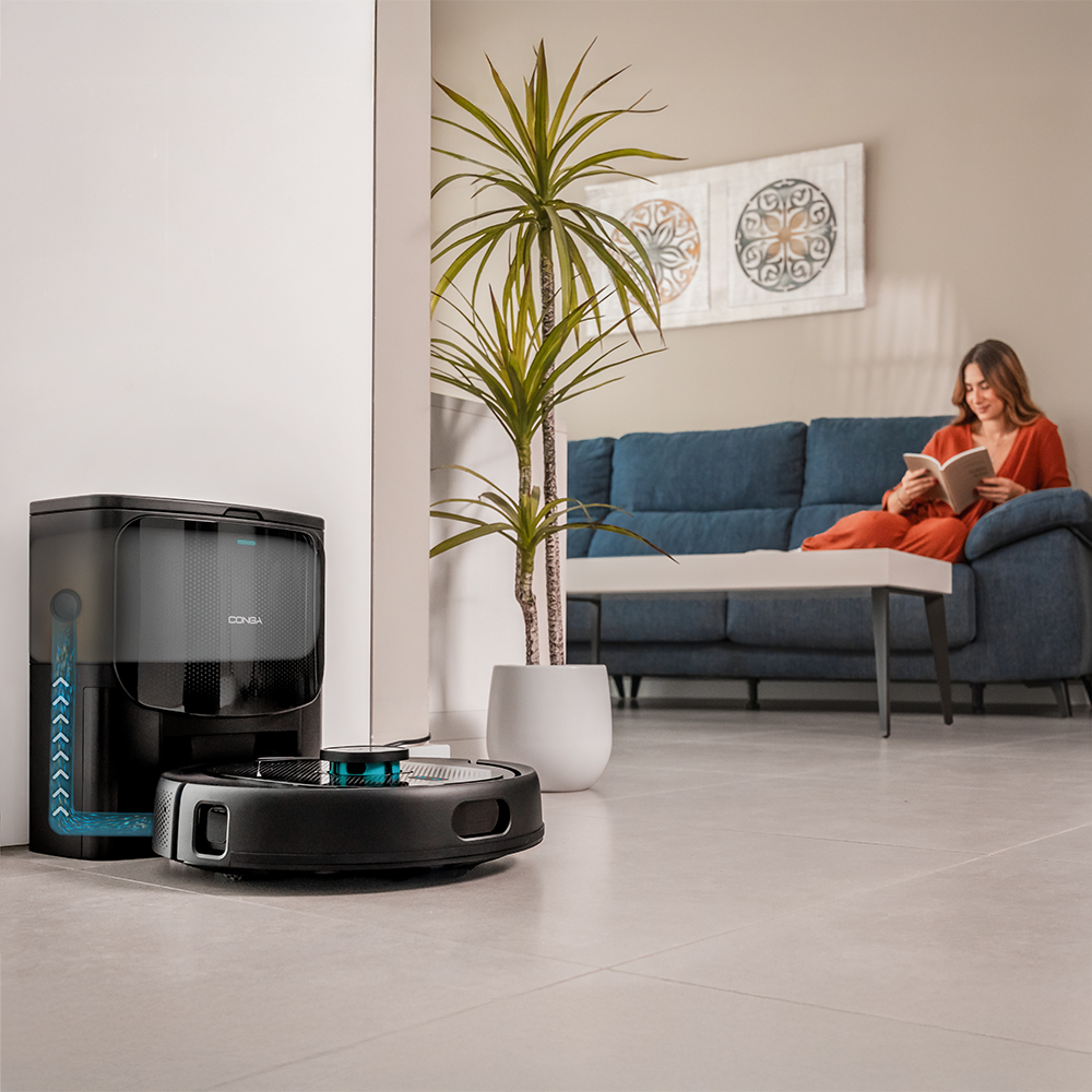 Una mujer lee en un sofá azul. Hay un robot aspirador en el suelo. Una planta grande en el medio.