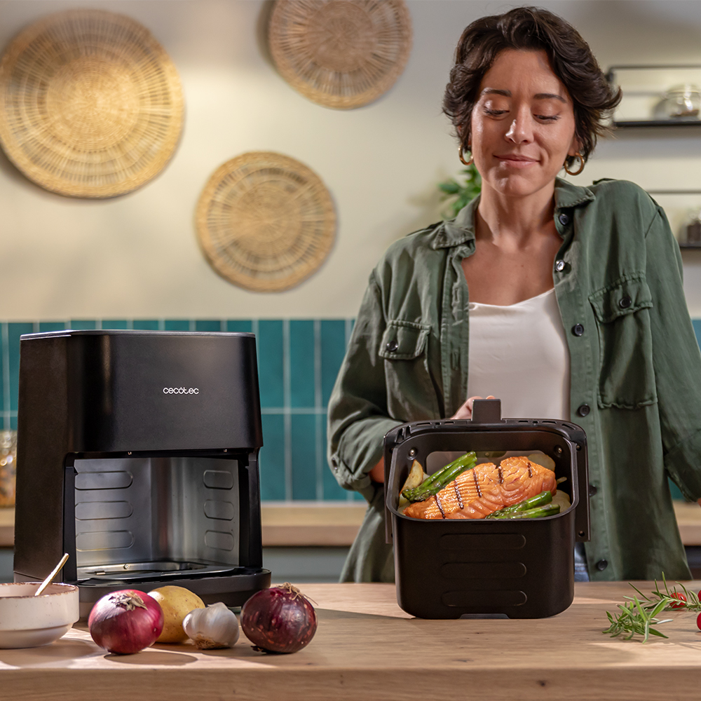 Mujer con freidora de aire, salmón y verduras. Cebollas y patatas en la mesa. Fondo de cocina con adornos de pared.