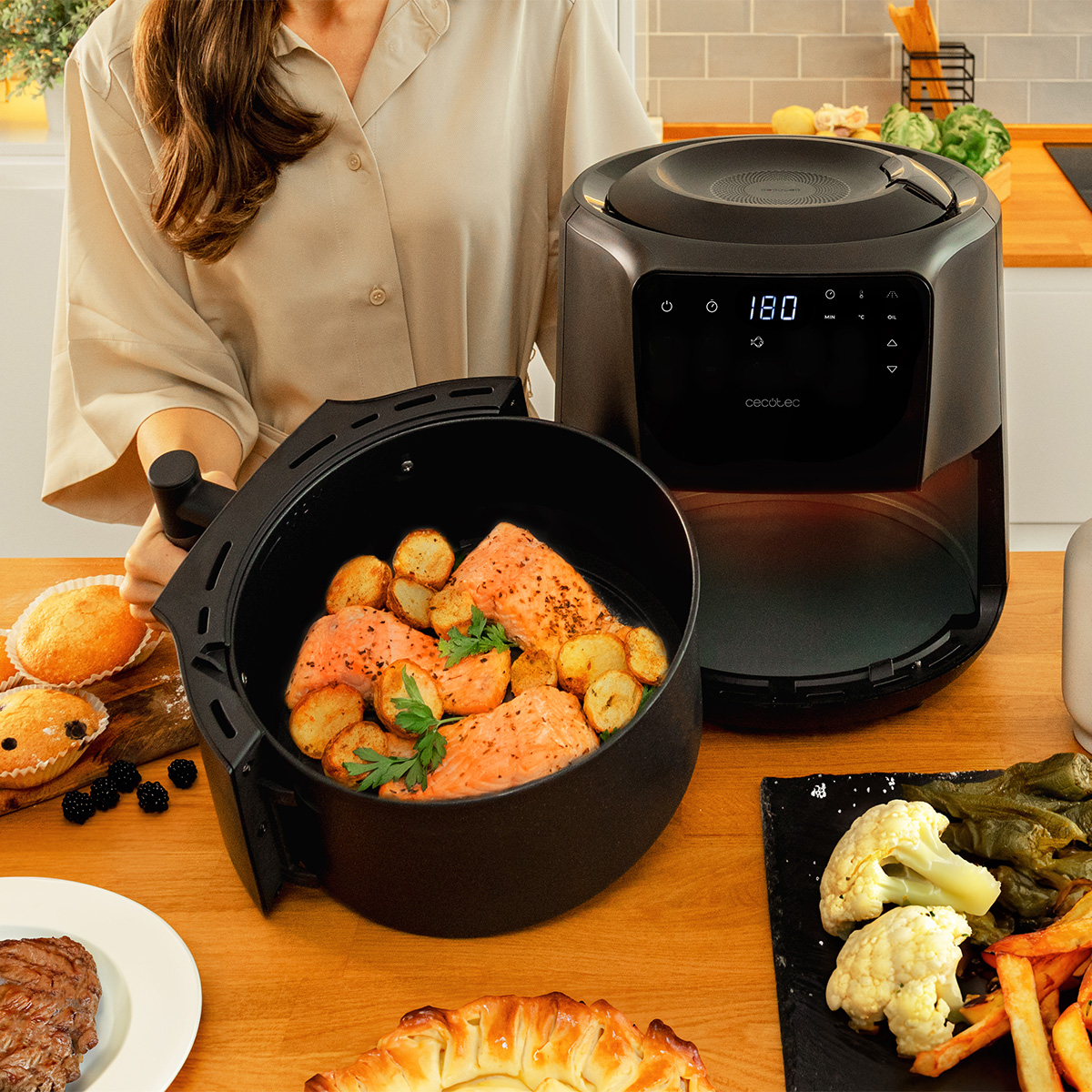 Mujer con freidora de aire. Salmón y patatas en la cesta. Otros alimentos incluyen bistec, magdalenas y verduras en una mesa de madera.