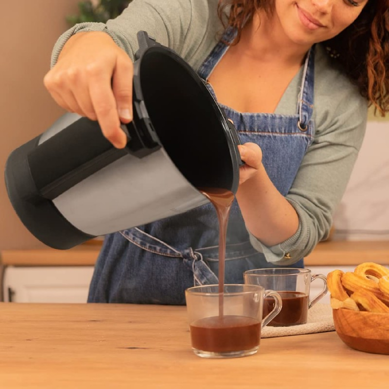 Mujer vertiendo chocolate caliente de una olla en tazas transparentes. Churros en un cuenco de madera sobre la mesa. Ambiente de cocina.