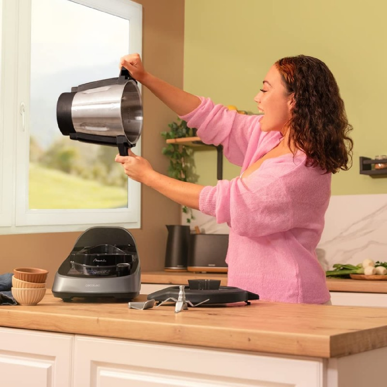 Mujer con suéter rosa sostiene un componente de electrodoméstico de cocina. Encimera con base de electrodoméstico y otros elementos. Ventana al fondo.