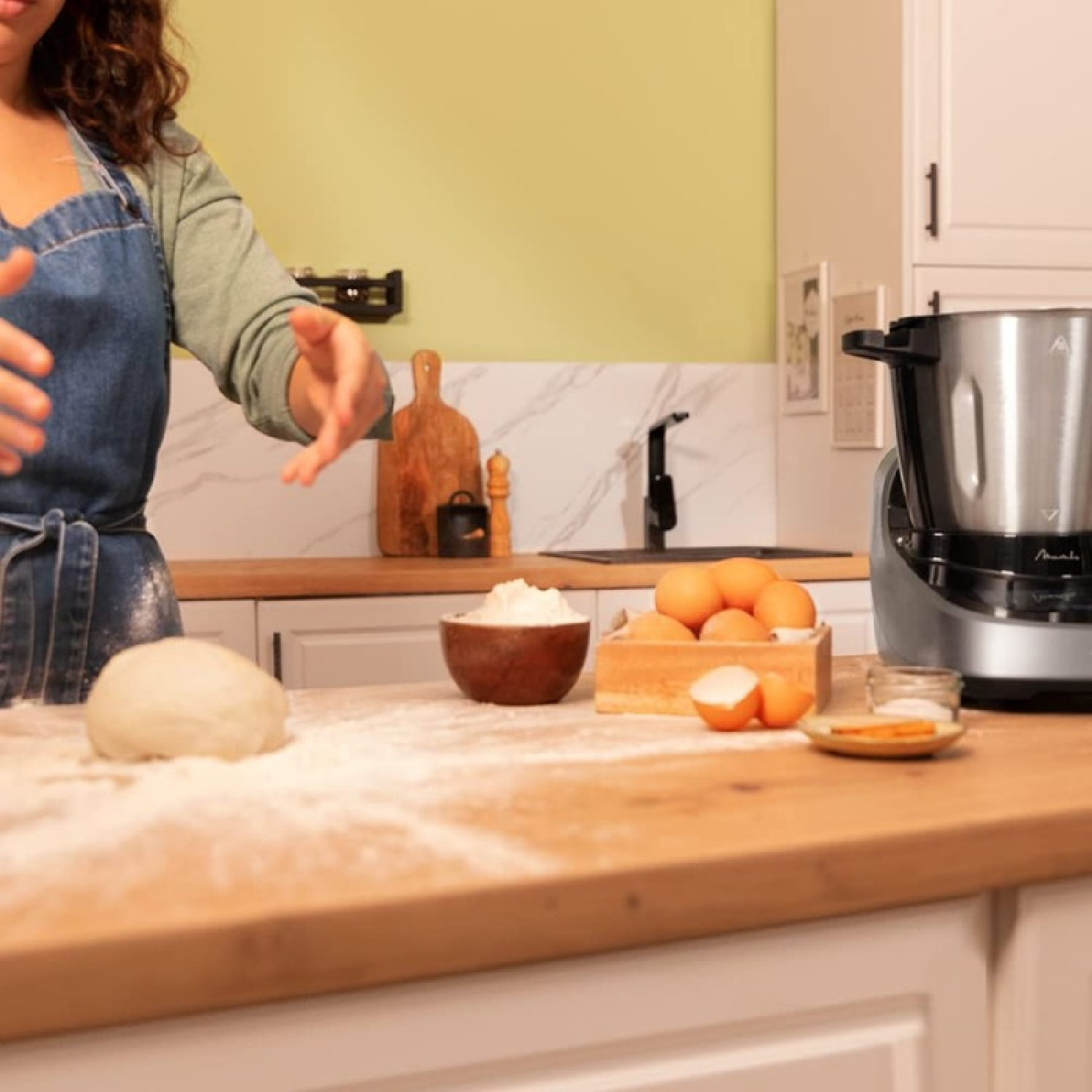 Una persona prepara masa en una cocina. Harina, huevos y una máquina de cocina están en la encimera de madera.