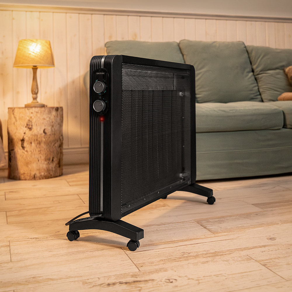 Calentador eléctrico negro sobre ruedas en una habitación. Una lámpara encendida y un sofá en el fondo. Suelo de madera.