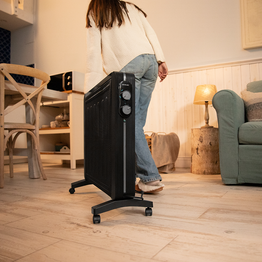 Una persona con jeans y un suéter blanco está de pie cerca de un calefactor negro sobre ruedas. En el fondo se ven un sofá, una lámpara y una mesa.