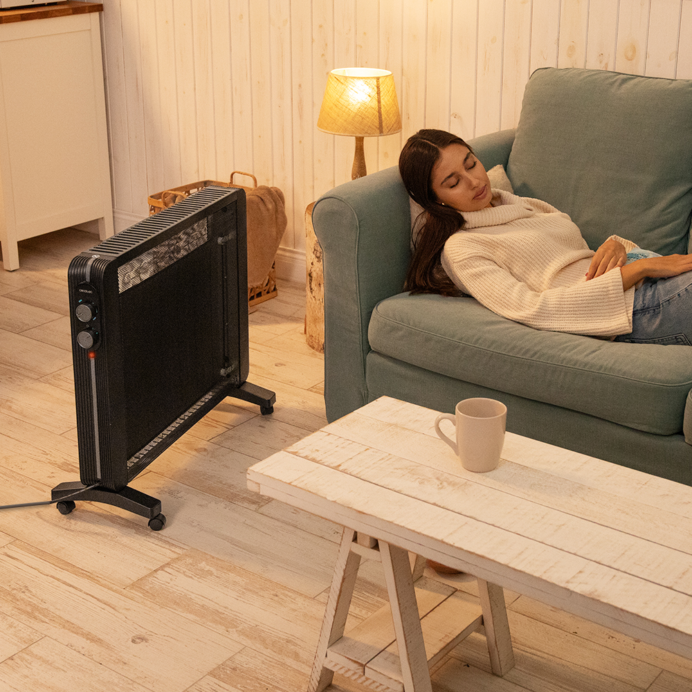 Mujer durmiendo en un sofá cerca de un calefactor. Una taza está en una mesa. Una lámpara está encendida y la habitación tiene pisos de madera.