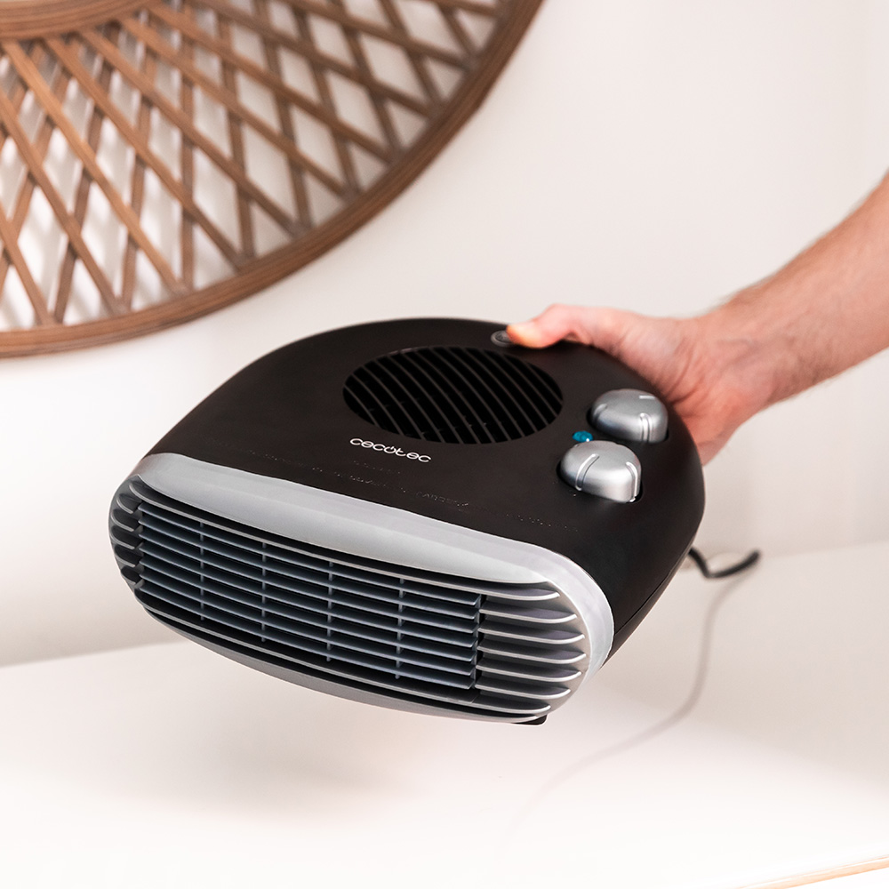 Una mano sostiene un calefactor eléctrico negro con detalles plateados. Tiene diales y una rejilla de ventilación. En el fondo, una pieza decorativa de madera en la pared.