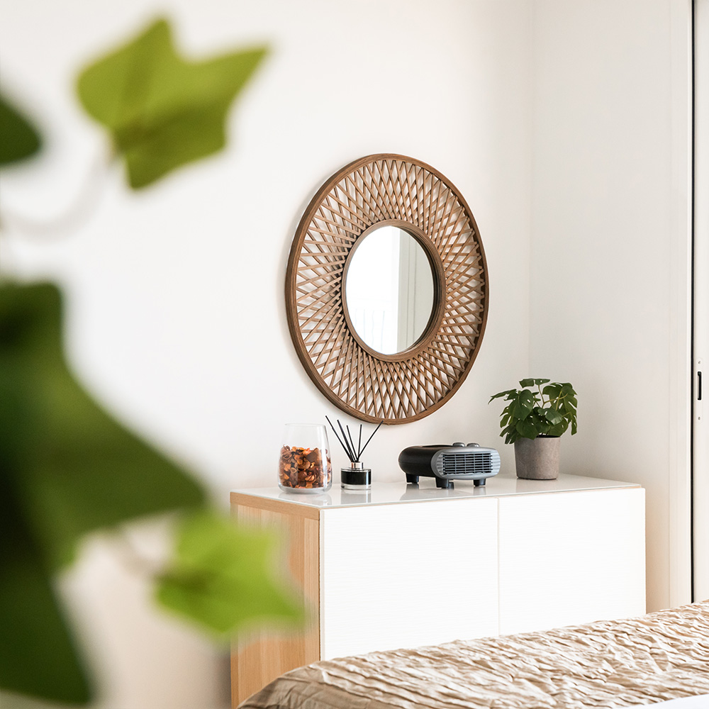 Escena de dormitorio con espejo redondo, armario blanco, planta y objetos decorativos. Una cama es visible en primer plano.