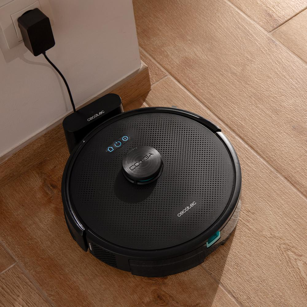 Una aspiradora robot Cecotec Conga negra con una base de carga en suelo de madera. La base está enchufada a una toma de corriente.