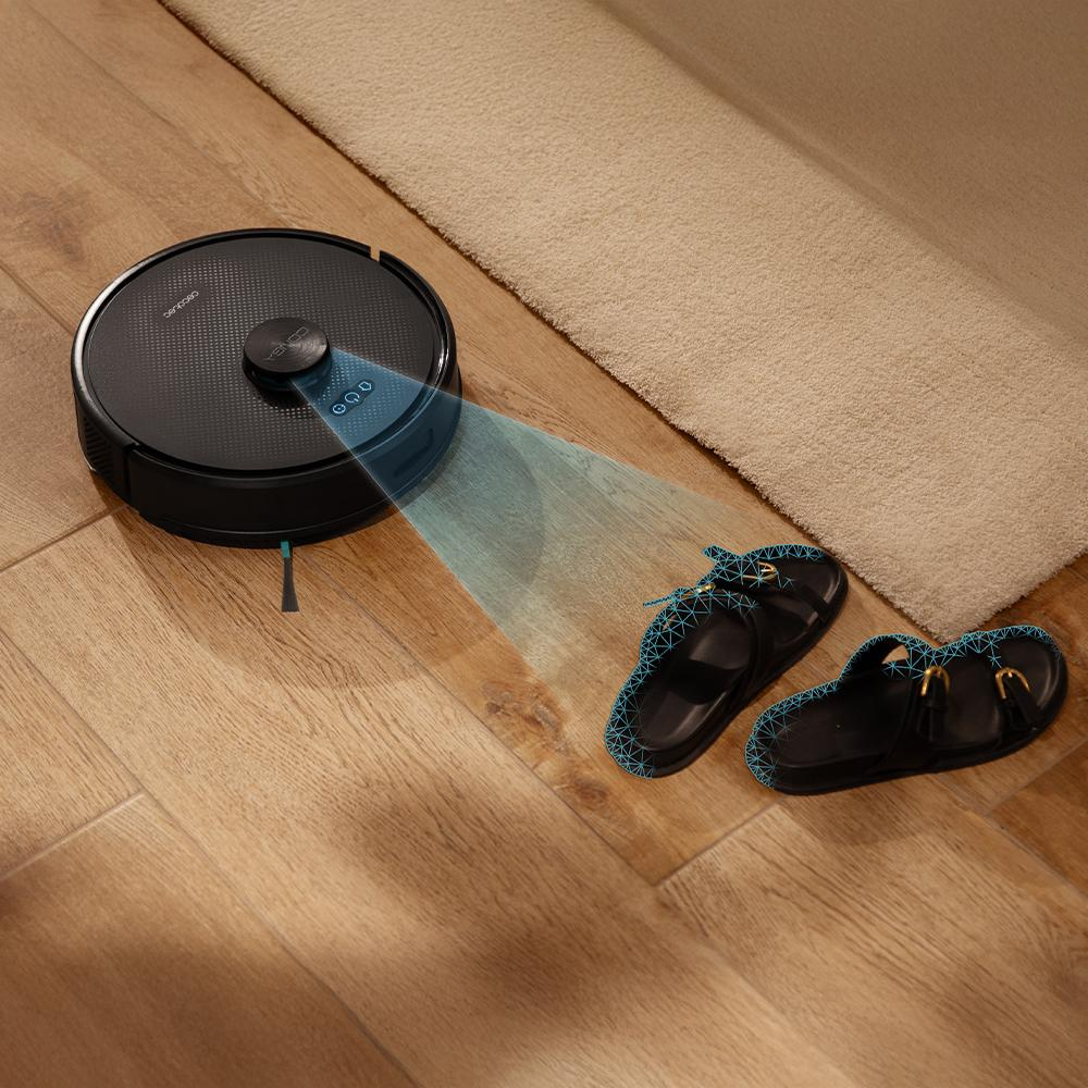 Una aspiradora robótica limpia un piso de madera. Hay sandalias y una alfombra cerca. La aspiradora tiene una proyección de luz azul.