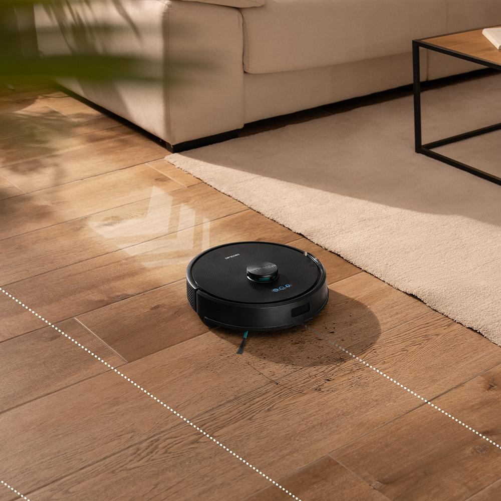 Una aspiradora robot negra en un suelo de madera. En el fondo, un sofá beige y una alfombra.