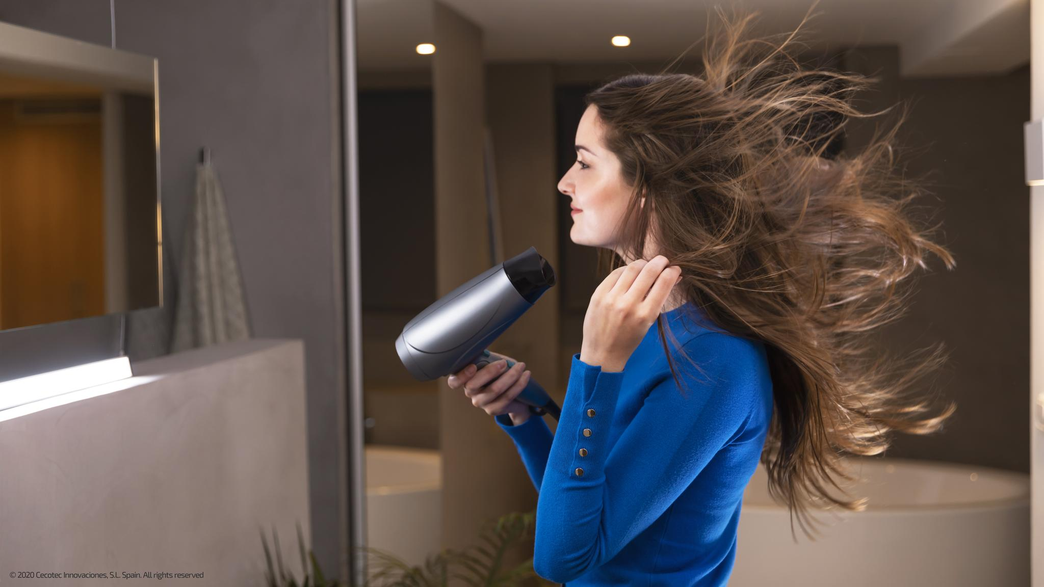 Mujer de azul secando el pelo con un secador en un baño. Pelo soplando. Espejo y luces visibles.