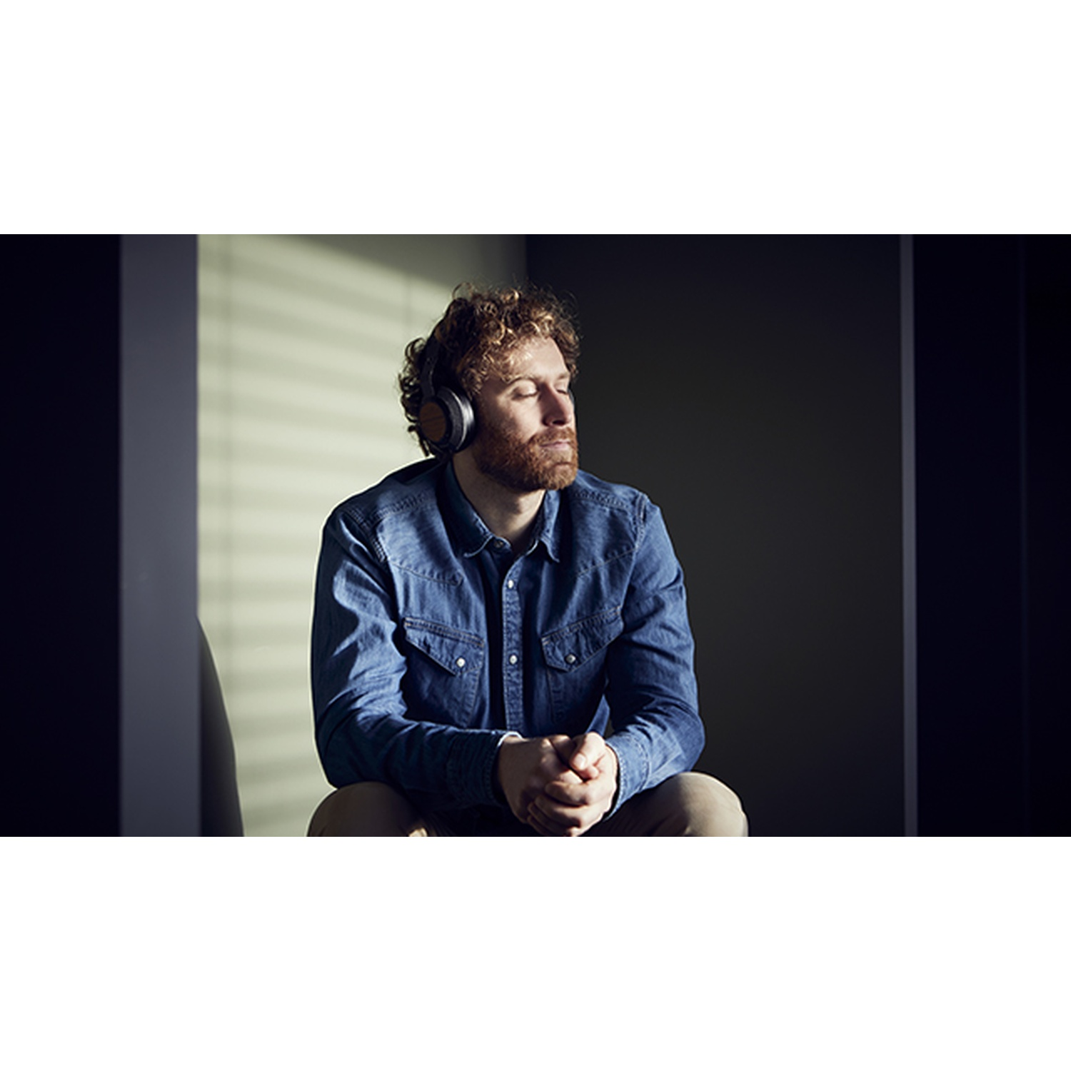 Un uomo con capelli rossi e barba con le cuffie è seduto con gli occhi chiusi. Indossa una camicia di jeans. La luce del sole attraversa il muro dietro di lui.