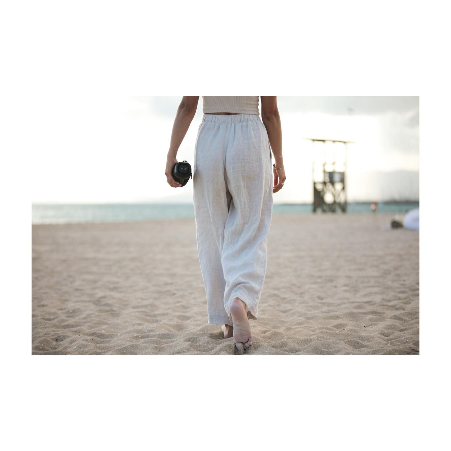 Una persona cammina su una spiaggia sabbiosa, tenendo in mano un oggetto nero. Indossa pantaloni bianchi e un top. Un posto di guardia è sullo sfondo.