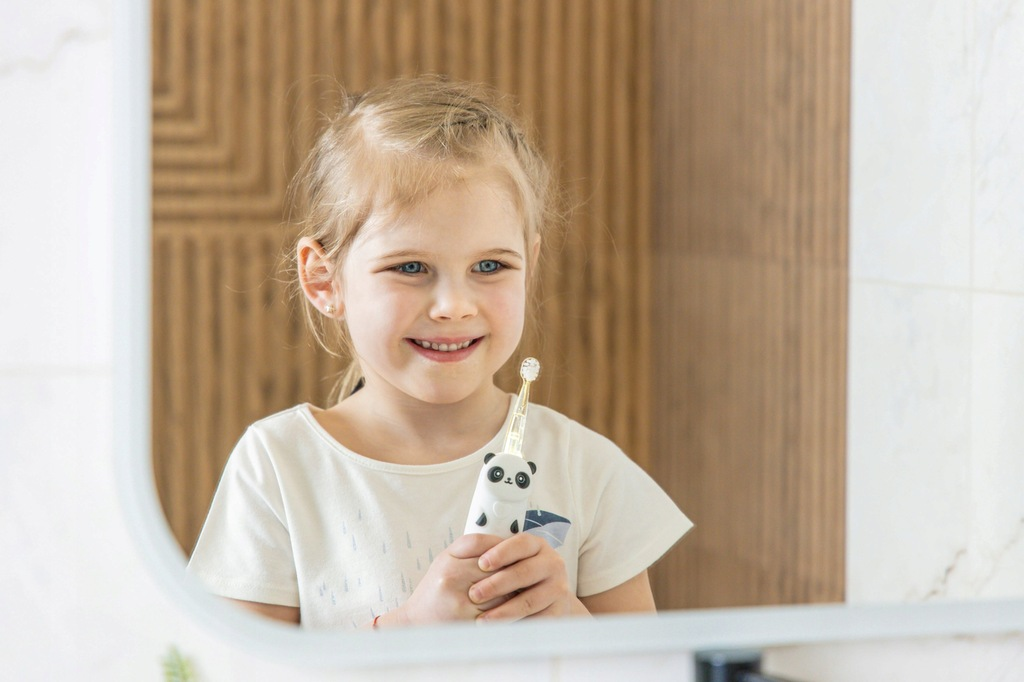 Uśmiechnięta dziewczynka trzyma szczoteczkę do zębów z pandą przed lustrem. Tłem jest drewniana ściana i marmurowe płytki.